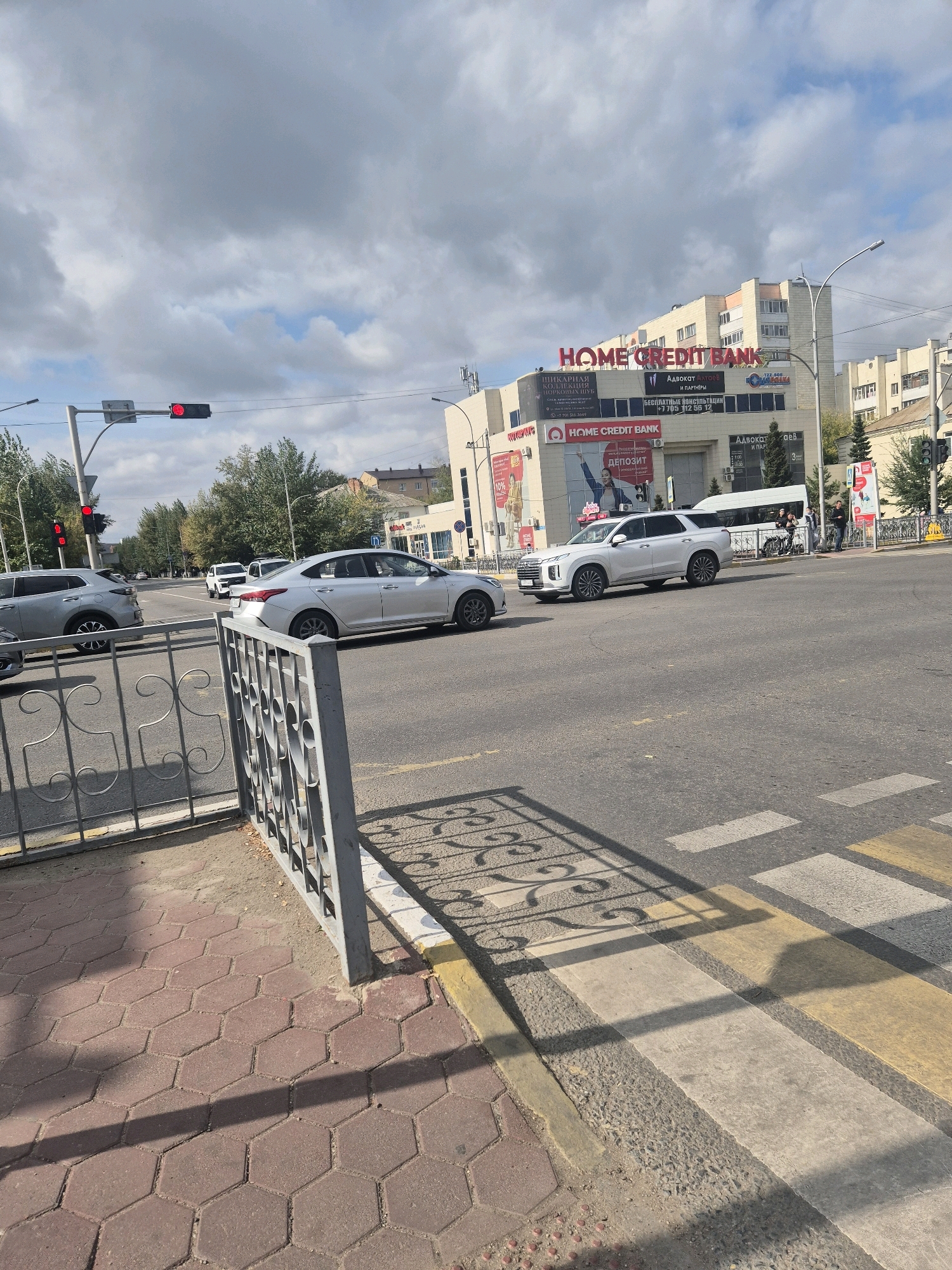 The image depicts a street scene in Kokshetau, Kazakhstan, on a cloudy day. A traffic light with a red signal is prominently displayed. Several cars are visible on the road, including a silver sedan and a white SUV. In the background, a building with a "HOME CREDIT BANK" sign is situated. A pedestrian crossing with yellow and white stripes is present on the road. A metal fence with decorative elements runs along the sidewalk, which is paved with hexagonal bricks. The environment appears to be urban, with trees and other buildings visible in the distance. There are a few people visible, including individuals standing near a bus and others potentially crossing the street.