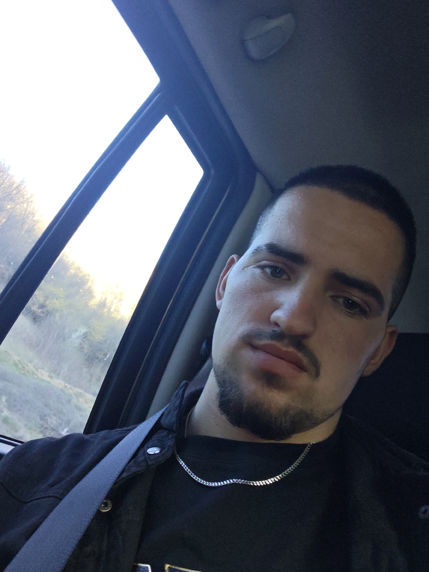 A man is in a car, presumably the driver. He is wearing a black shirt, a black jacket, and a silver chain. He has dark hair and a beard. He is wearing a seatbelt. The view outside the car window is blurry but appears to be a landscape with trees.
It is not possible to determine the city of Devin, Bulgaria based on this image alone.