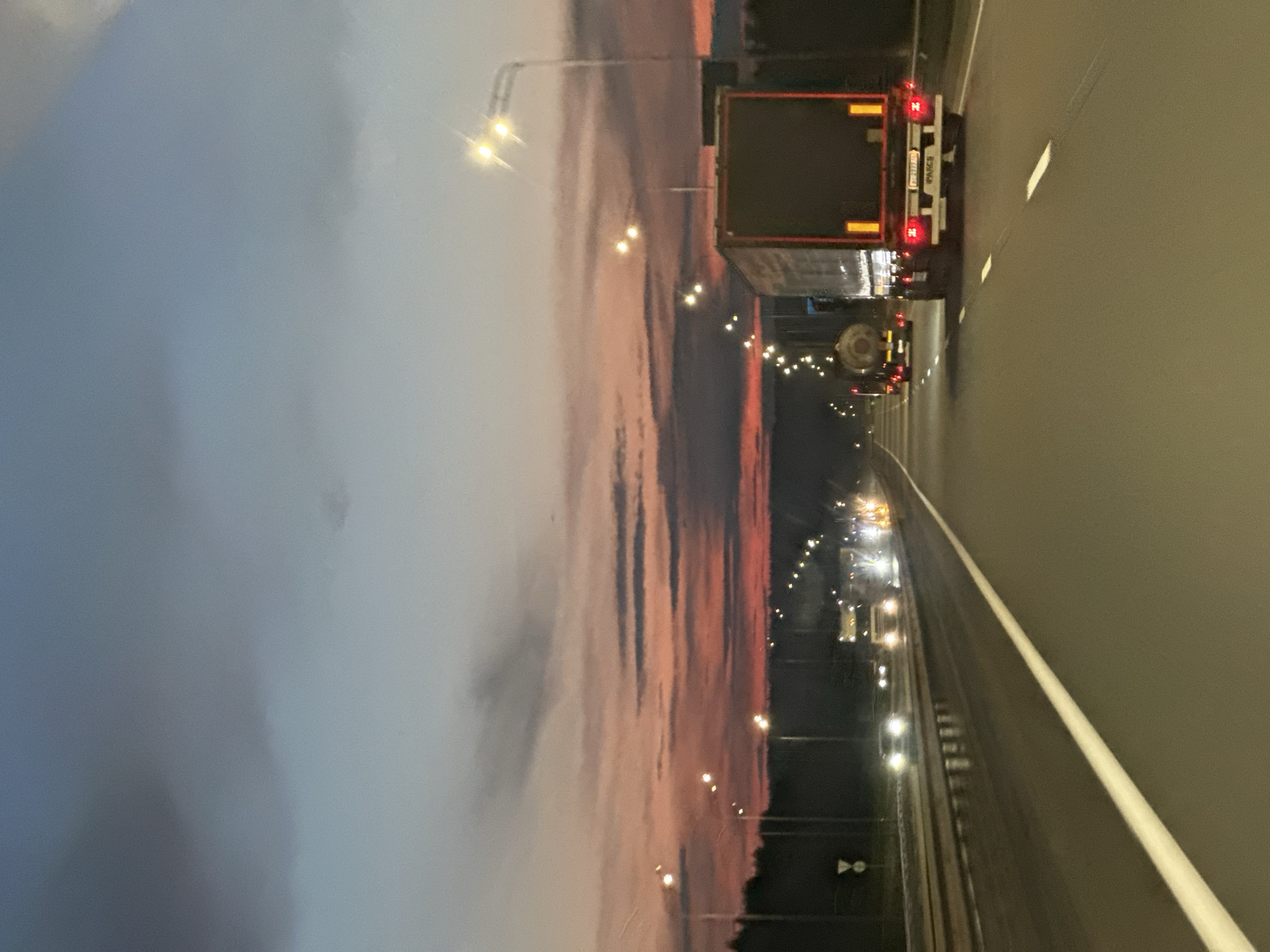 The image shows a highway at dusk. The sky is a gradient of blue and pink, with clouds casting shadows. There are several trucks on the road, including one in the foreground with a dark trailer and red taillights. Streetlights illuminate the highway. The road is paved and has white lane markings. A line of trees is visible on the left side of the road. The location is identified as the city of Zvanka, Russia.