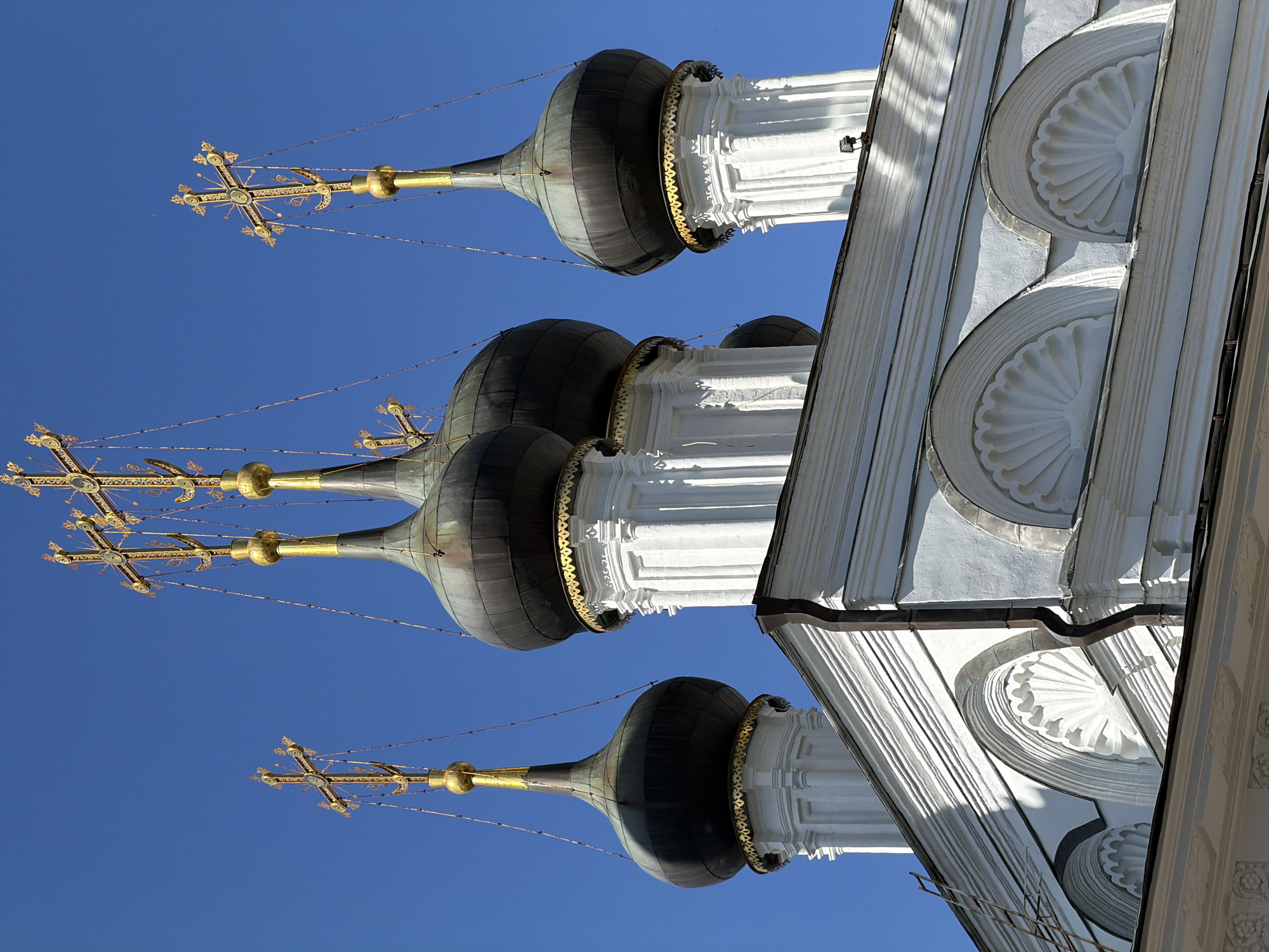 The image shows a close-up, low-angle view of the domes and crosses of the Church of the Annunciation in the Kremlin, Moscow, Russia. The church features multiple onion domes, dark in color with golden crosses atop each spire. The domes are supported by white, fluted pillars, and the architecture exhibits ornate detailing with decorative gold trim around the base of each dome. The building's facade is white, with arched decorative elements and visible cornices. The sky is a clear, bright blue, indicating a sunny day. No people are visible in the image. The overall scene depicts a section of a historical religious building against a cloudless sky.