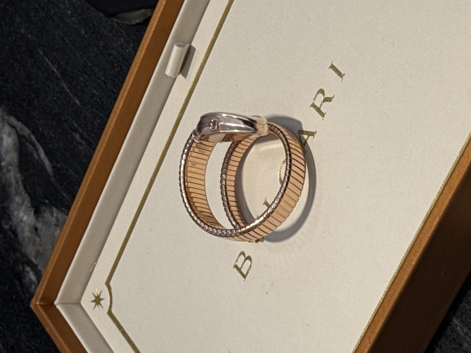 A rose gold Bvlgari ring is presented in an open, light-colored box with gold trim and a star emblem. The ring is coiled and features a watch face. The box sits on a dark, textured surface, possibly a table, in what appears to be a retail or personal setting in Tel Aviv, Israel. No people are present in the image.