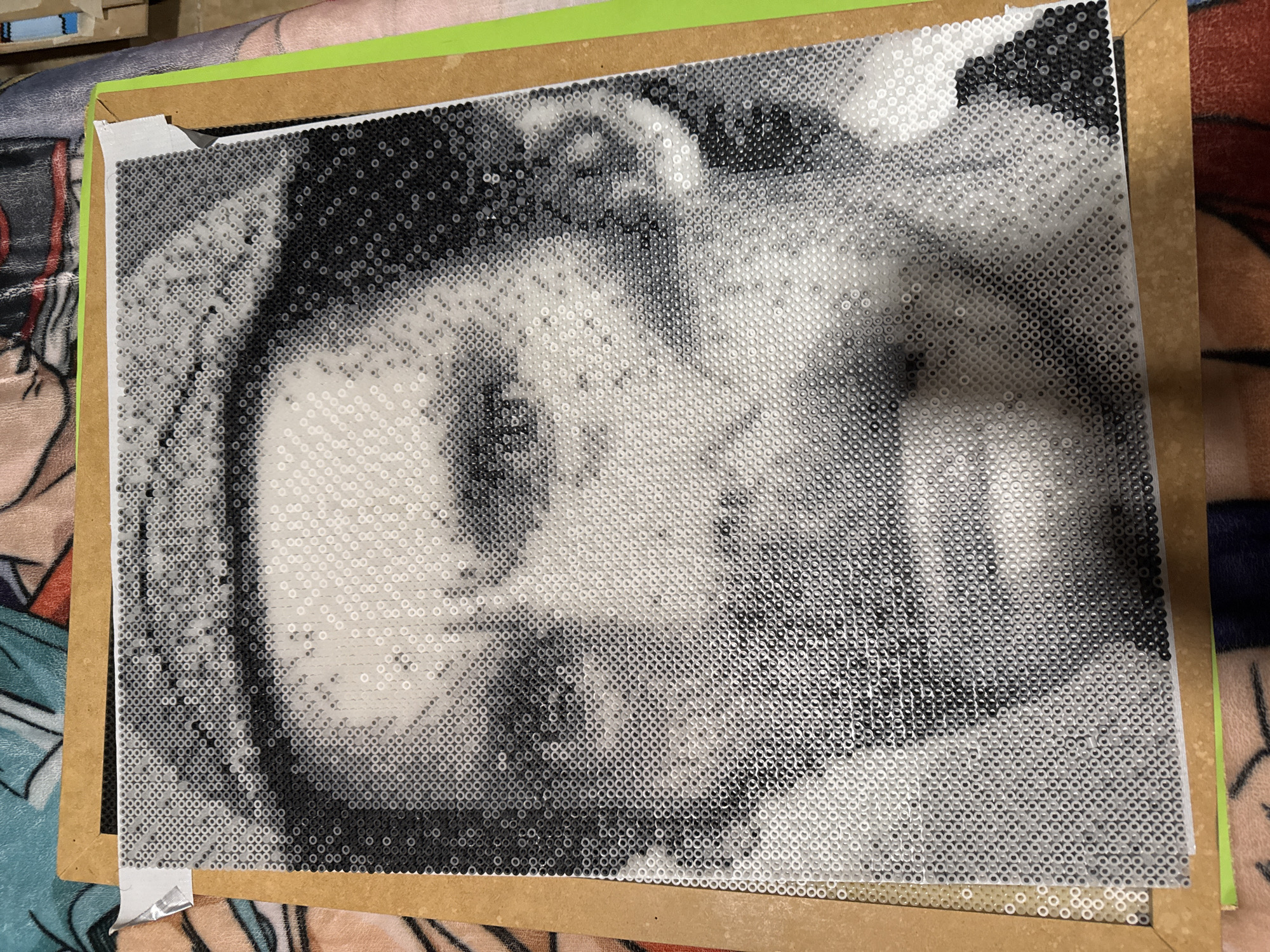 This media file features a grayscale portrait of a person, meticulously crafted from countless small, circular Hama or Perler beads. The beads, in shades of black, white, and various greys, are arranged in a grid on a white pegboard to form the facial features, including eyes, nose, and mouth, creating a distinct pixelated effect. The expression of the subject appears neutral or contemplative.
The finished bead art is mounted or rested upon a light brown wooden or cardboard backing, with white tape visible at the corners, securing the bead boards. A thin green border runs along one edge of the backing. The entire piece is positioned on a soft, patterned fabric surface, possibly a blanket or bedspread, which is visible in the background and foreground, out of focus. This indicates an indoor, domestic setting for the photograph, taken in Puebla, Mexico. No visible text is present in the image. The artwork itself is the central subject, representing a completed craft project.