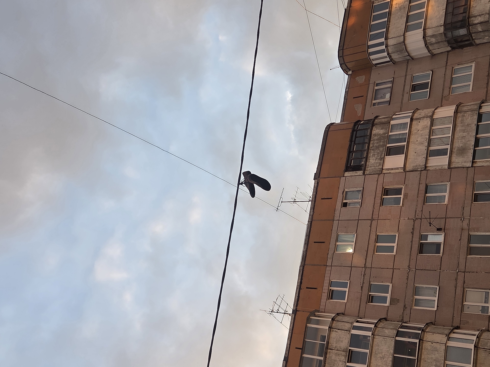 The image depicts a view looking up at a pair of shoes hanging from a utility line against the backdrop of a cloudy sky and the side of a multi-story apartment building. The building is of Soviet-era construction, with a grid-like facade featuring numerous windows. There are some antennas. The sky is overcast with hints of blue and orange near the horizon. The location is Perm, Russia. There are no visible people in the frame.
