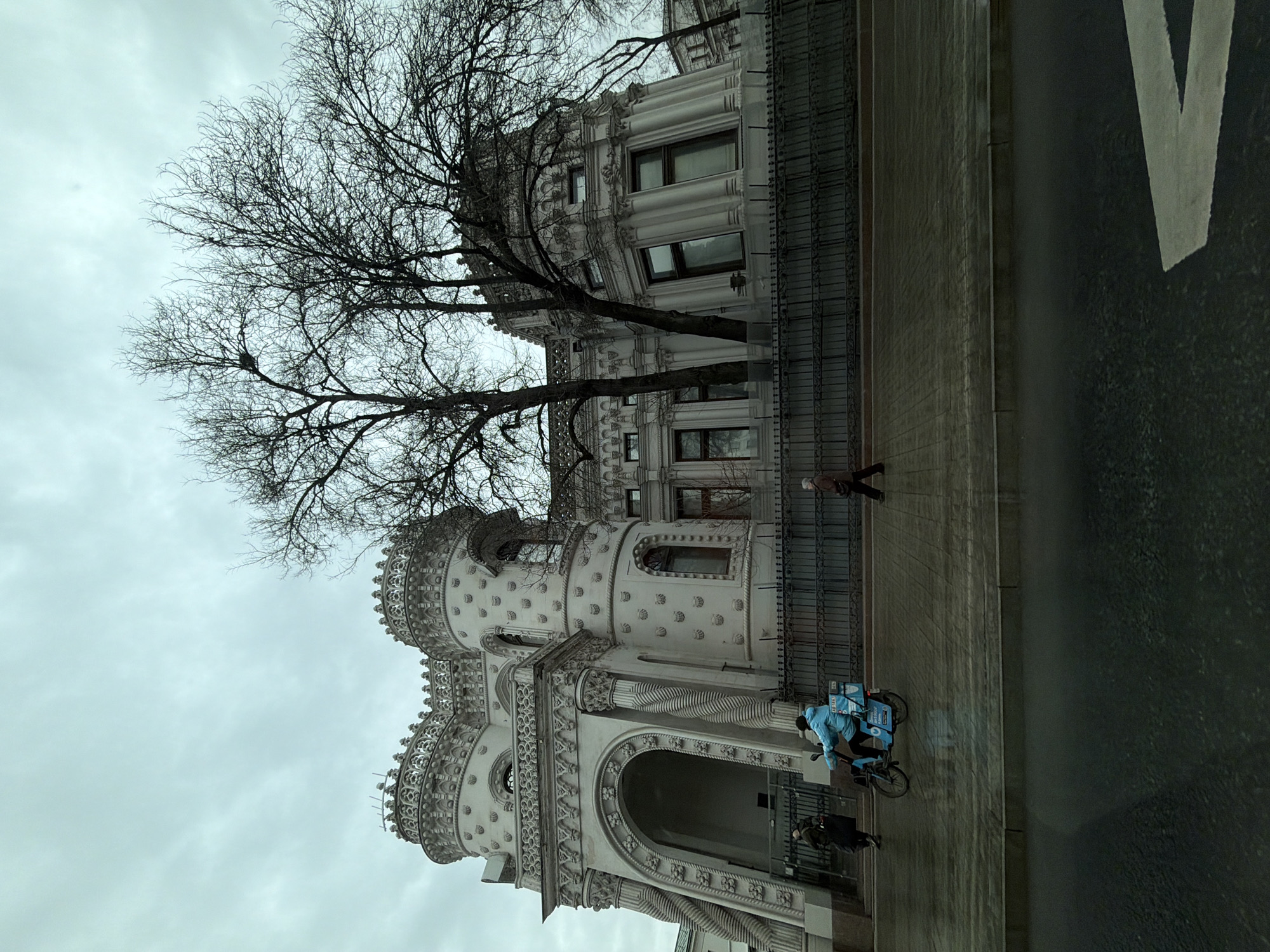 The image displays the Morozov Mansion, located at Vozdvizhenka Street, 16, Moscow, Russia, under an overcast sky. The multi-story building features a light-colored, ornate facade extensively decorated with numerous repetitive shell-like patterns. Two prominent, round towers with intricate, crenellated tops are visible, alongside a large arched entrance supported by spirally twisted columns. An ornate, dark metal fence runs along the property's perimeter in front of the mansion, behind which several bare deciduous trees stand, indicating a late autumn or winter season.
The street and sidewalk in the foreground are visibly wet, suggesting recent precipitation. Three individuals are present on the sidewalk: one person in dark clothing walks away from the building's main entrance, while another person, wearing a light blue jacket, operates a light blue electric bicycle equipped with a rear delivery box, moving from left to right. A third pedestrian, dressed in a brown coat, walks from right to left further down the sidewalk. A partial white directional arrow is painted on the wet asphalt road surface in the lower right foreground.