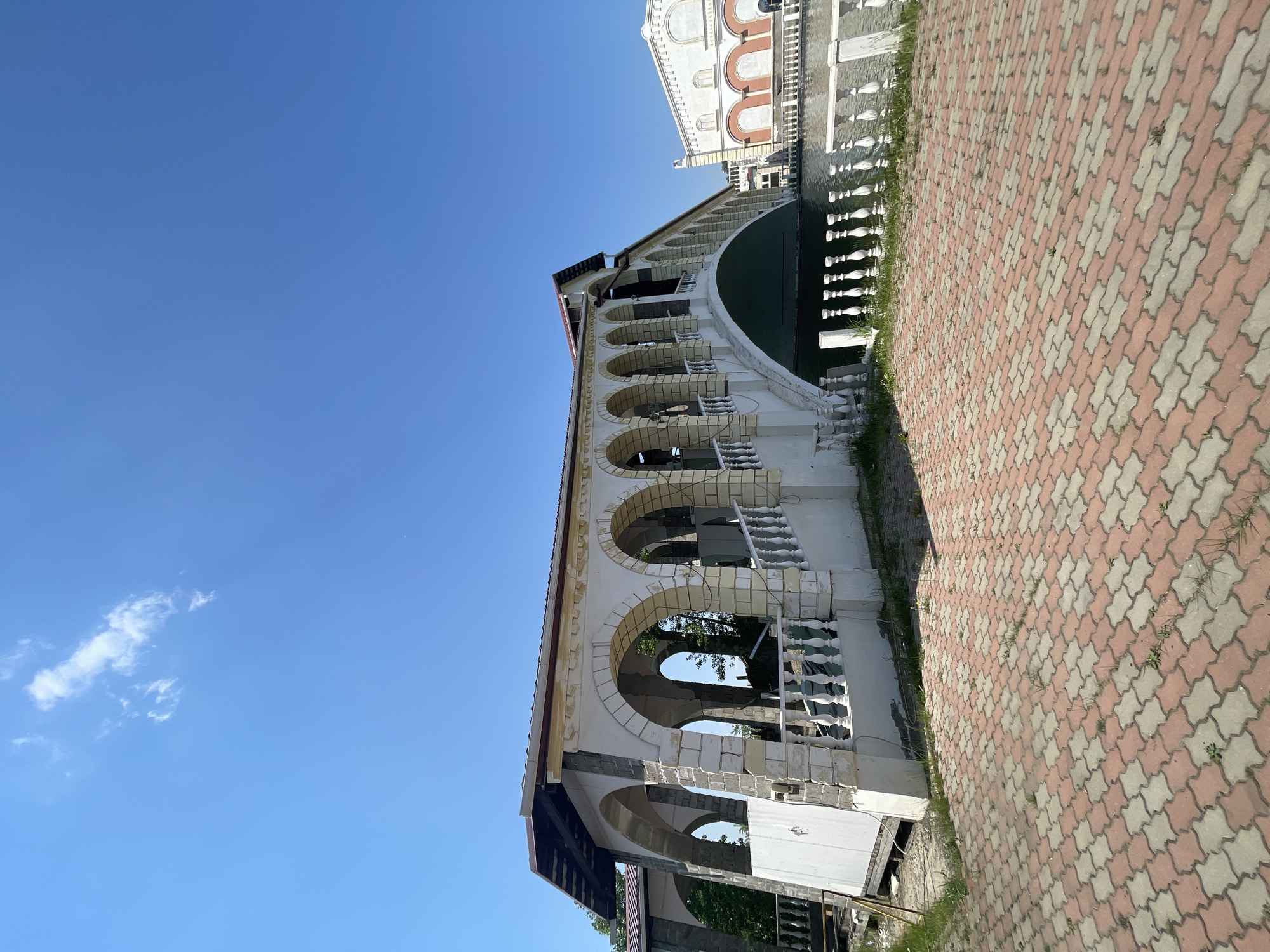An outdoor scene captured on a clear day under a bright blue sky with minimal wispy clouds. The dominant feature is a partially constructed or renovated bridge-like structure, exhibiting classical architectural elements such as arches and white balustrades. This structure, primarily white with sections revealing grey building material, spans over a dark body of water visible beneath and to its right. The roofline displays exposed wooden beams and dark red trim. In the immediate foreground, a patterned paving of interlocking reddish-brown and light beige tiles is visible, with sparse weeds growing in the crevices. To the right, beyond the water, a large, ornate white building with prominent arched recesses and decorative facade elements stands in the background. No individuals or specific actions are depicted. The location is in the city of Unknown, Russia.