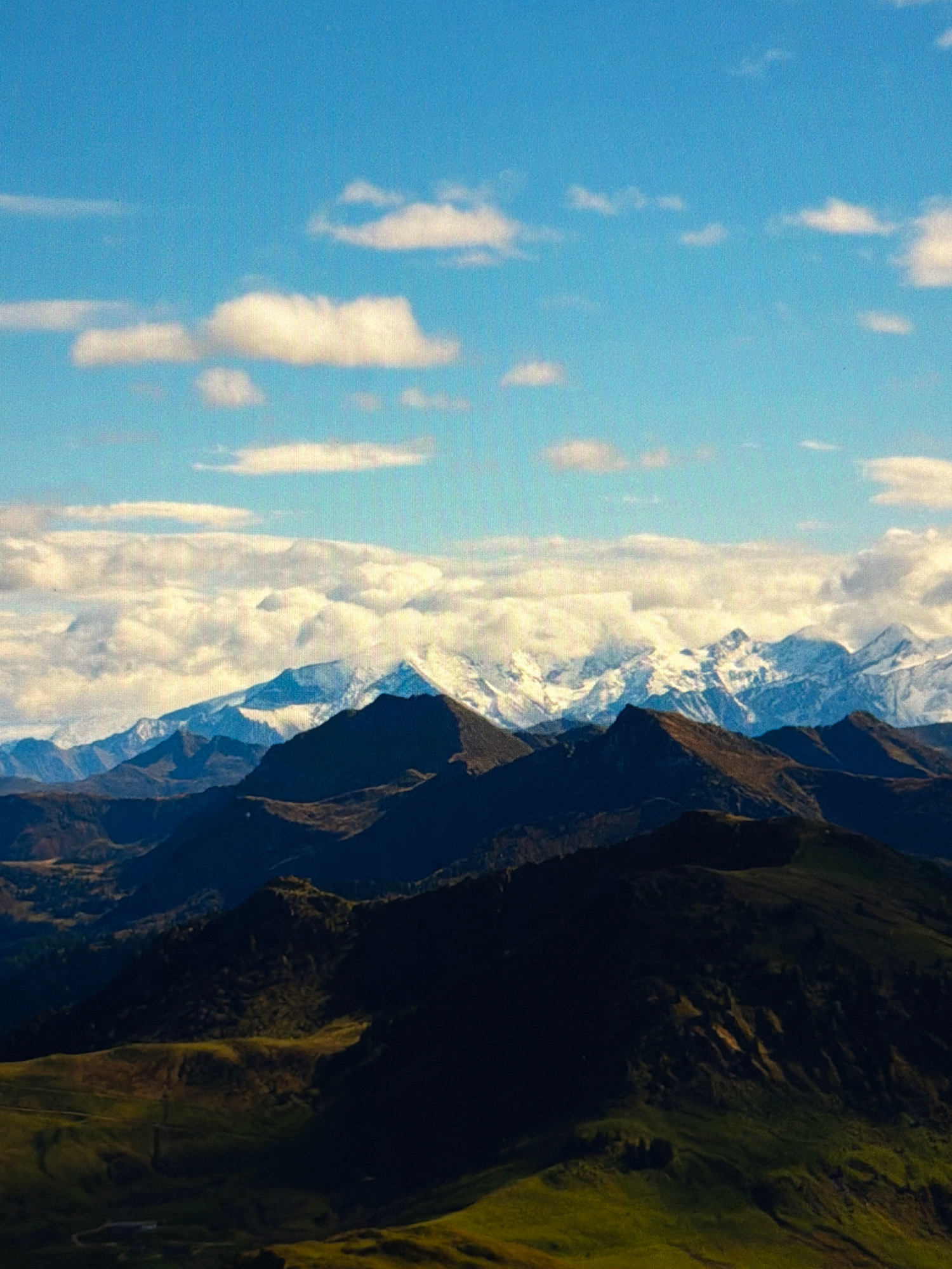 This is a landscape photograph of a mountain range under a blue sky with scattered clouds. The mountains in the foreground are dark and shadowed, while the distant peaks are snow-capped and illuminated. The sky is a clear blue with a few white clouds. There are no people or man-made objects visible in the image. The image does not depict the city of Kafr Yasif, Israel, as it shows a natural mountainous environment.