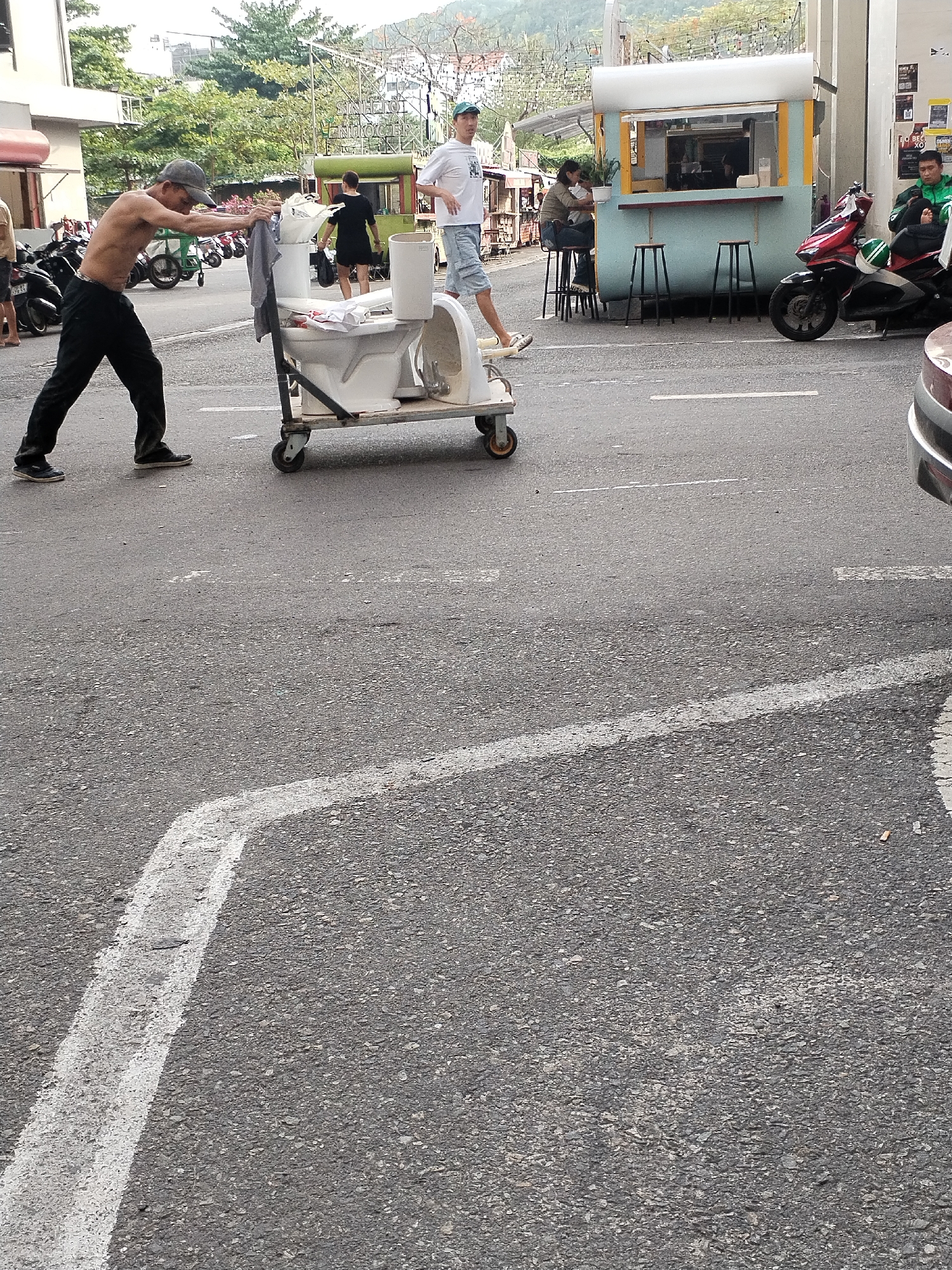 In an urban setting, likely Bac Nha Trang Ward, Vietnam, a man pushes a handcart laden with two white toilets down an asphalt street. The man, shirtless and wearing black pants and a baseball cap, leans forward as he propels the cart from the left side of the frame. One toilet is upright on the cart, while the other lies on its side, with some clothes draped over the cart's handle.
Several other individuals are present in the background. To the right of the man and the cart, another man in a white t-shirt and shorts walks away from the viewer. Further back and to the right, a small light blue food stall with a yellow trim is visible, featuring two stools where a person is seated. Next to the stall, a red scooter is parked, and a person in a green jacket, possibly a delivery rider, is seated on it.
The street itself is paved with asphalt and features faded white lines. Parked motorbikes are visible on the left side of the street, indicating a commercial or residential area. Buildings and trees line the background. The lighting suggests it is daytime, possibly overcast, with no harsh shadows.
Visible text includes a partially obscured sign in the distance that reads "SINGLETON PRODUCTORS" and a poster near the food stall with the text "O BEE XO". The overall scene captures a slice of daily urban life, with people going about their business amidst the practical movement of goods.