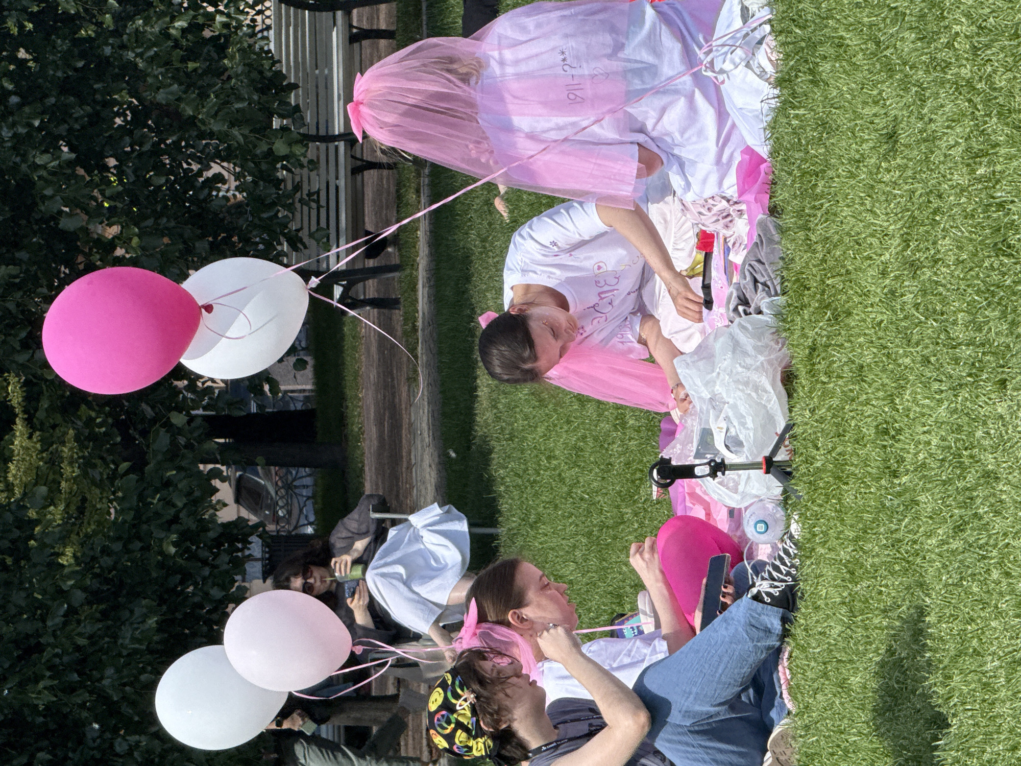 In Saint Petersburg, Russia, on a sunny day, four young women are having a picnic on a lush green lawn. Two women in the foreground are seated on a pink blanket, engaged in an activity. The woman on the left wears a dark t-shirt and blue jeans, with a colorful bandana on her head. The woman next to her, wearing a white t-shirt and a pink veil, is focused on something in her hands. To their right, another woman, also in a white t-shirt and a pink veil, is similarly occupied. In the background, a woman sits in a chair, holding a phone and a green drink. Pink and white balloons are scattered around the scene, adding a festive atmosphere. The background reveals trees and benches, suggesting a park setting within the city.