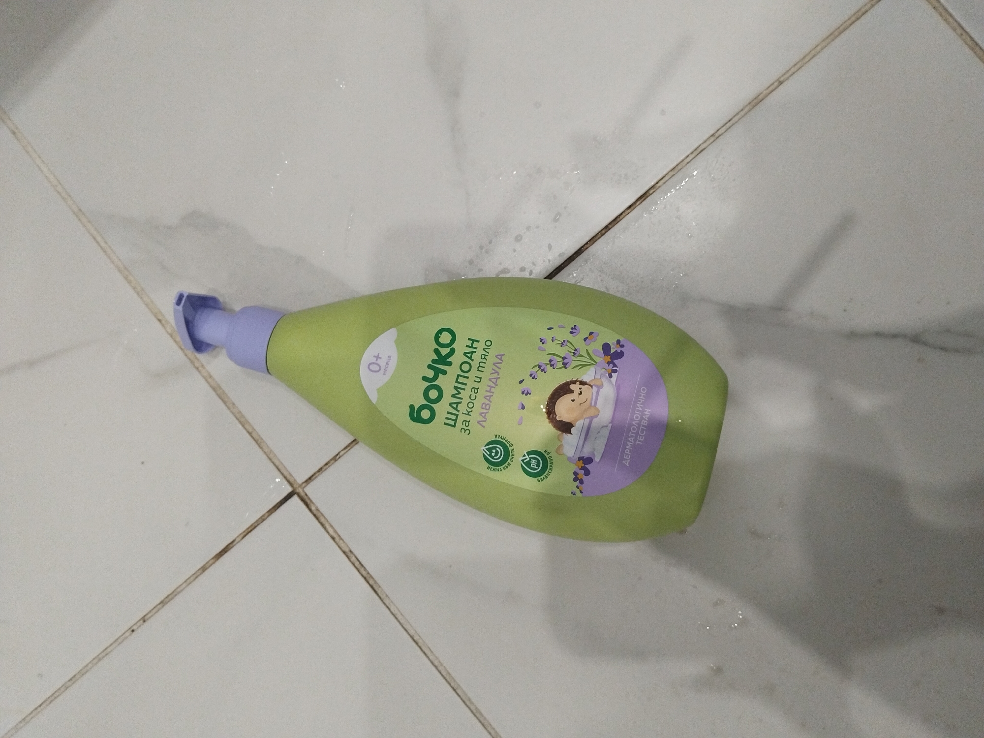A bottle of "Бочко" brand baby shampoo lies on a white tiled surface. The bottle is green with a purple pump and features Bulgarian text, an illustration of a sleeping child with lavender, and labels such as "0+ месеца" and "PH БАЛАНСИРАНО." The tiles appear to be in a bathroom or similar space, and the scene suggests the bottle is lying on the floor. The overall environment is clean but slightly wet, as indicated by water droplets on the tiles. There are no specific clues in the image to place it in Stara Zagora, Bulgaria, beyond the possible presence of Bulgarian text on the shampoo bottle.
