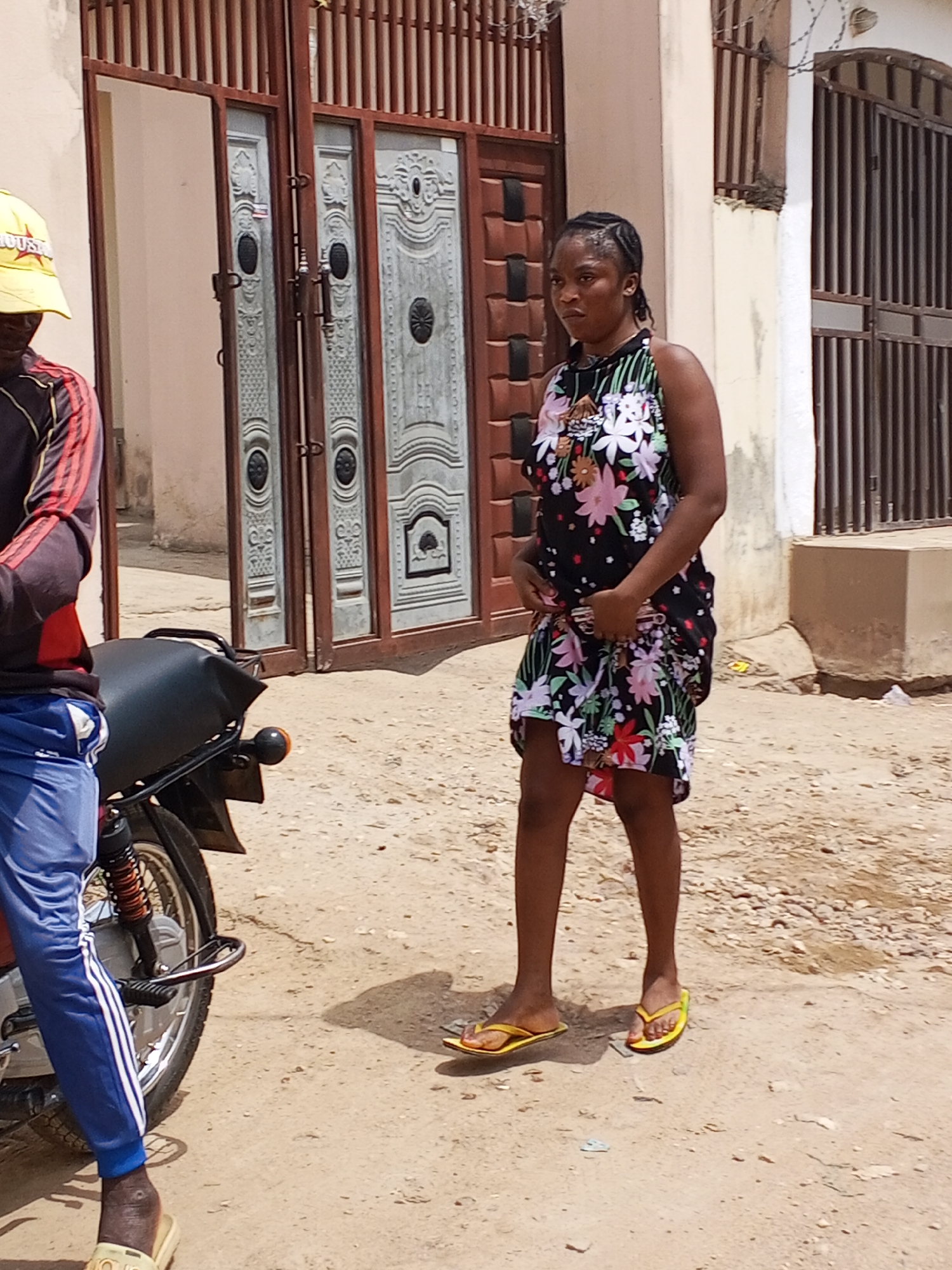 A young woman stands on a dirt road in Adonkolo, Nigeria. She wears a floral halter-neck dress and yellow flip-flops. Her hair is braided. To her left, a man wearing blue track pants with white stripes, a black and red long-sleeved shirt, and a yellow hat sits on a motorcycle. Behind them, is a large gate in front of a building. The road is sandy and uneven.