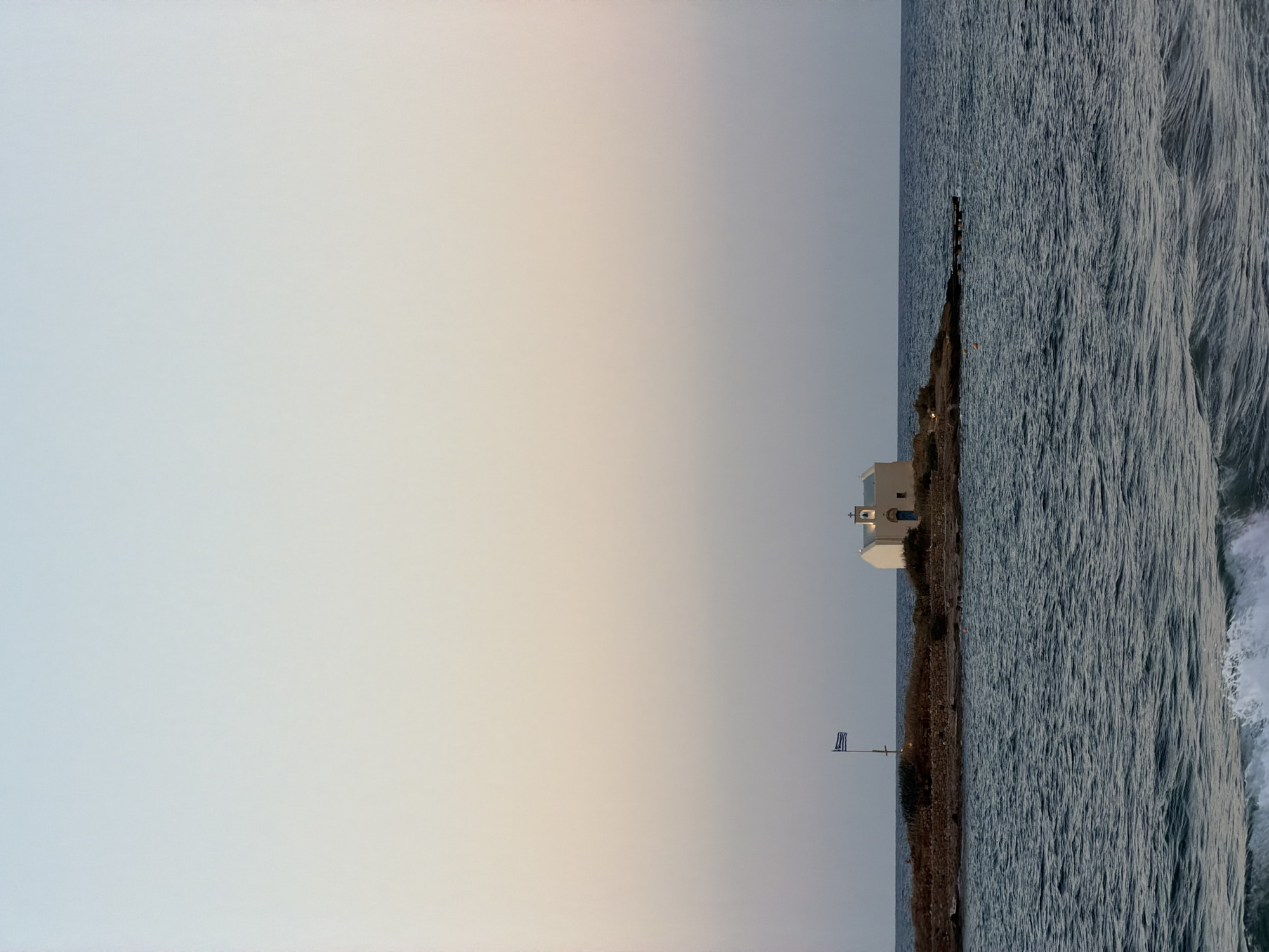 The image depicts a small, white chapel situated on a rocky island in the sea. A Greek flag flies on a flagpole to the left of the chapel. The chapel has a blue door and a small bell tower. The sea is a deep blue with gentle waves, and there is a hint of a sandy beach in the foreground. The sky is a soft gradient of pale blue and peach, suggesting either sunrise or sunset. The scene is serene and picturesque, with no people visible. The location is the island of Agios Nikolaos, off the coast of Malia, Greece.
