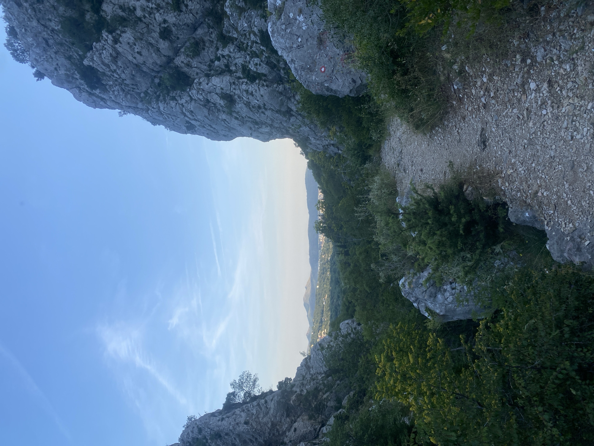 The image presents a scenic view of a rocky, mountainous landscape, likely a hiking trail, captured during the late afternoon or early evening. The perspective is looking forward along a path that leads towards a distant valley and hills.
In the foreground and midground, large, grey, rugged rock formations dominate both sides of the scene, creating a natural frame. These rocks are interspersed with abundant green vegetation, including dense bushes and scattered trees, suggesting a verdant, Mediterranean-like environment. To the right, a narrow, unpaved trail composed of dirt and gravel winds into the distance. A clear red and white circular trail marker, indicating a hiking route, is painted on a large rock next to the path on the right.
Beyond the immediate rocky outcrops and vegetation, the landscape opens up to reveal a wide valley or plain. In the far distance, faint outlines of mountains or hills are visible, and amidst them, several small white structures suggest a remote settlement or town.
The sky above is a clear, pale blue, transitioning to a lighter hue near the horizon, with delicate, wispy white clouds adding texture. The lighting is soft and diffuse, characteristic of golden hour, casting gentle shadows and highlighting the natural textures of the rocks and foliage. No people are visible, emphasizing the tranquility and unspoiled nature of the location. The scene evokes a sense of peaceful exploration and natural beauty in a rugged, remote setting.