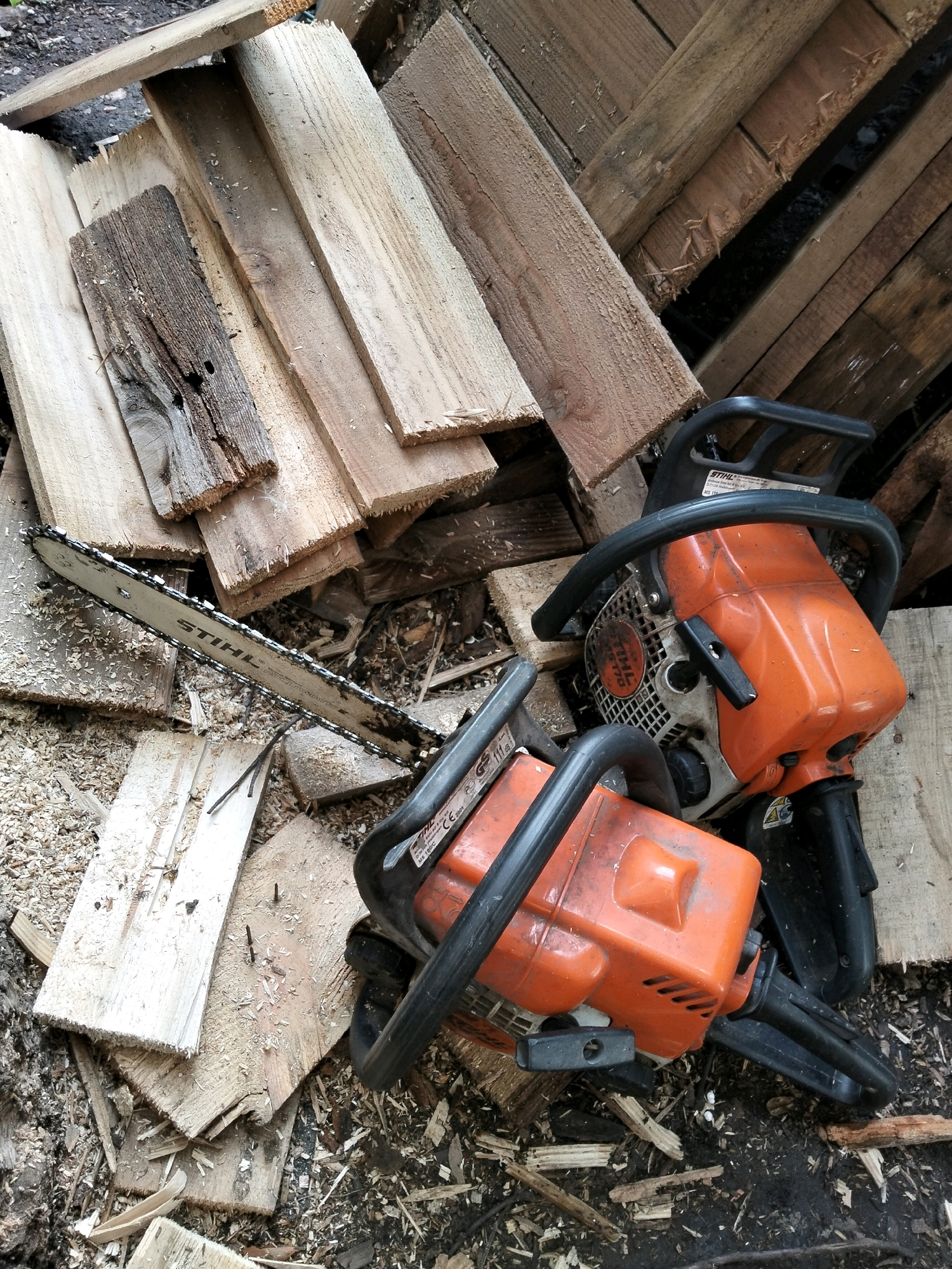 The image shows two orange and black Stihl chainsaws on a pile of wood scraps. The chainsaws are in the foreground, with the one on the left partially obscured by the one on the right. The wood scraps appear to be a mixture of rough-cut lumber and sawdust. The background is a jumble of larger wooden planks and debris. The scene is outdoors, likely in a backyard or workshop area. The lighting suggests it is daytime. The specific location in Oktyabrsky, Russia, cannot be determined from the image alone.