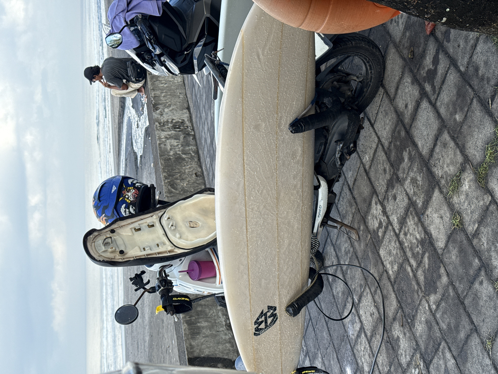 The scene, seemingly in Unknown, Unknown, shows two motorbikes parked on a patterned grey stone walkway near a beach. A surfboard is strapped to one of the motorbikes. The seat of the white motorbike is open, revealing a purple cup inside. A blue helmet sits on the seat. A man in a dark shirt and pants is sitting at the edge of the stone walkway looking at the beach and ocean. Waves are visible on the beach. The sky is cloudy.