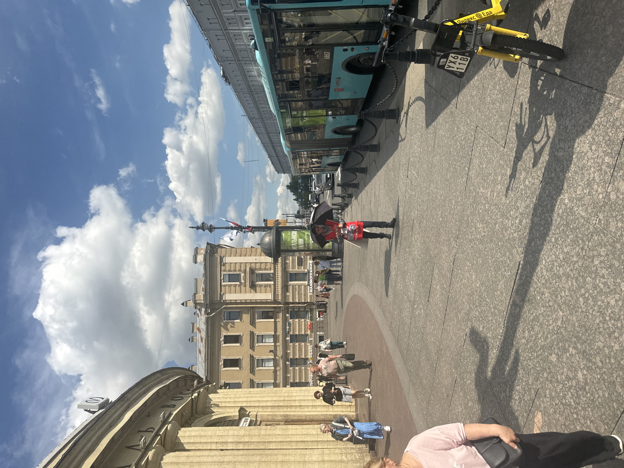 The image depicts a street scene in Saint Petersburg, Russia, likely near a metro station given the signage. In the foreground, on the left, a woman wearing a pink shirt and black pants is walking away from the camera, carrying a black purse. The ground is paved with granite tiles. Further down the walkway, several people are walking in various directions. A man in a red vest and black pants, holding a black umbrella, walks towards the viewer. To the right, a long, teal-colored bus is parked along the curb. A yellow electric scooter with license plate YX6 11B is parked in front of the bus. Behind the bus, a multi-story building with numerous windows lines the street, under a bright blue sky with scattered white clouds. The architecture suggests a historical urban setting.