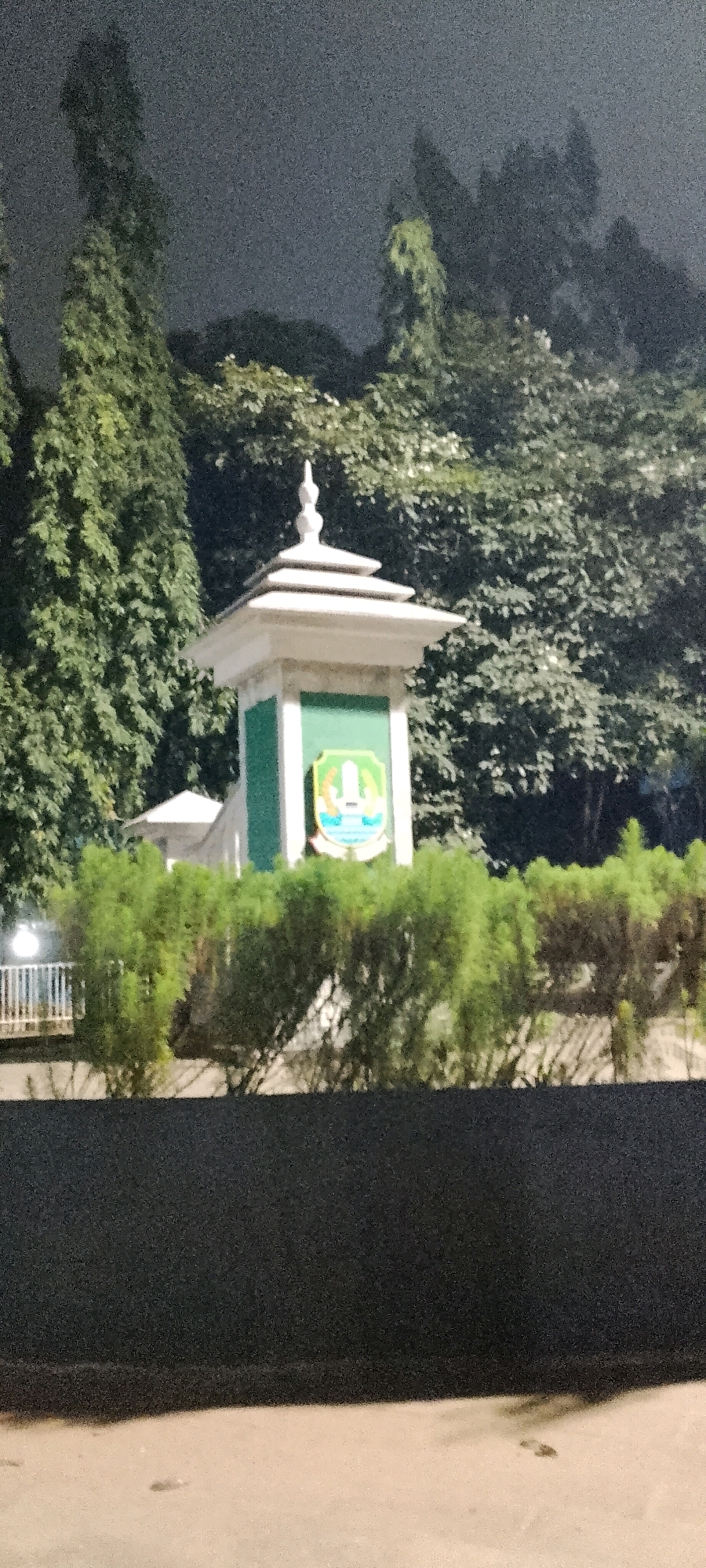 The image shows a monument at night, possibly a landmark or a gate, with a distinctive white, tiered roof. The monument is decorated with a green panel featuring a crest or emblem. Behind the monument, there are lush green trees and foliage, indicating a park or a well-maintained outdoor area. In the foreground, there are manicured bushes and a dark, solid surface that could be a wall or a raised platform. The overall atmosphere is serene, and the lighting suggests it is nighttime. This monument appears to be located at the gate of the city of Bekasi, Indonesia, given its prominence and decorative elements.