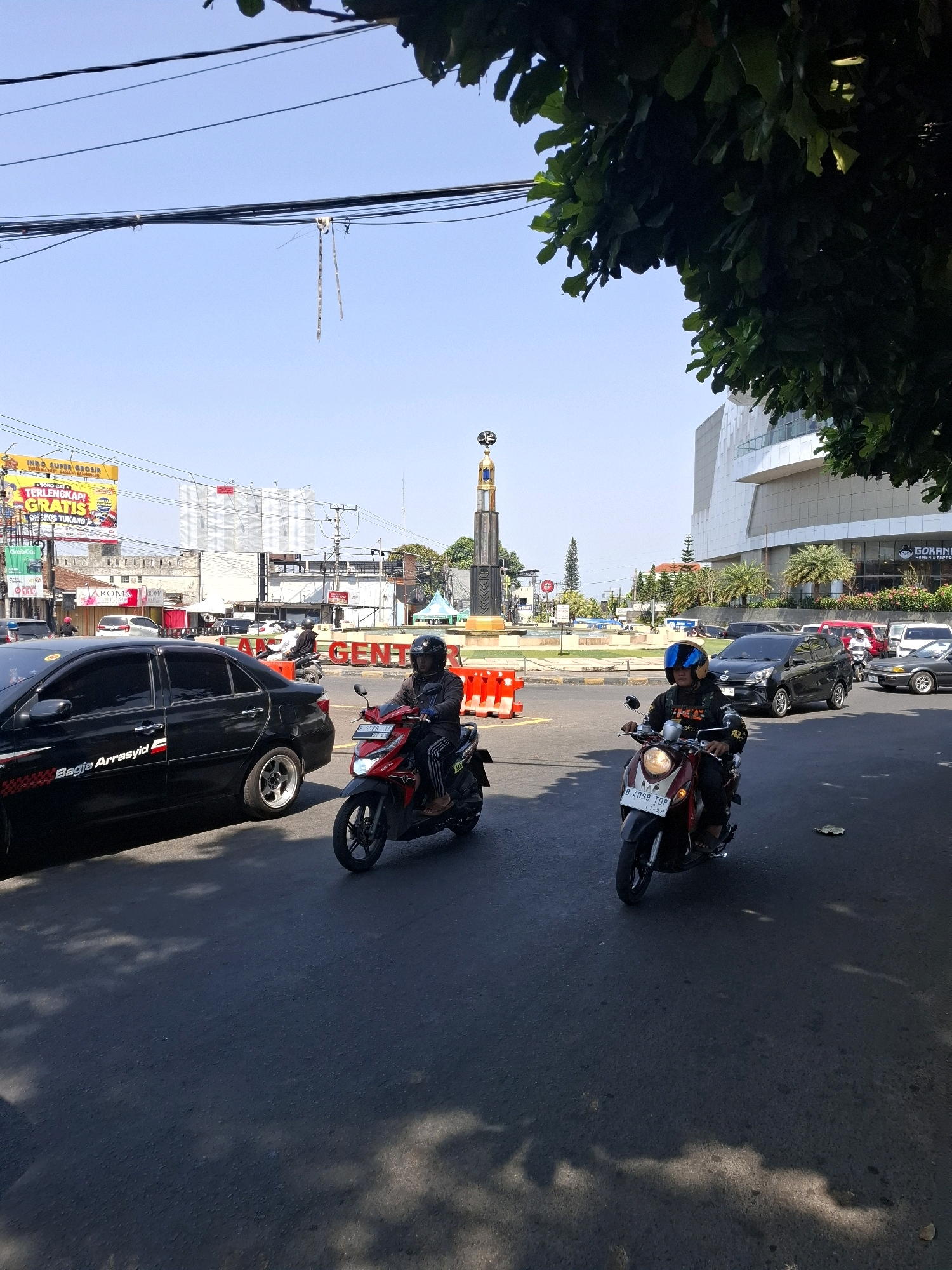 A street scene in Cianjur, Indonesia, on a sunny day. In the foreground, two individuals are riding motorcycles. The motorcycle on the left is red and driven by a person wearing a helmet and dark clothing. The motorcycle on the right is also red and is ridden by a person wearing a helmet and a dark jacket. A black sedan with the text "Bargia Arrasyid" on its side is in the left foreground. In the middle ground, traffic flows around a roundabout with a tall monument featuring a gold finial. Orange traffic barriers are visible. A large, modern building with a white facade and balconies is in the background on the right, with palm trees in front of it. Several other cars and motorcycles are on the road. Overhead, power lines crisscross the clear blue sky, and the leaves of a large tree are visible in the upper right corner. A billboard is visible in the distance on the left.