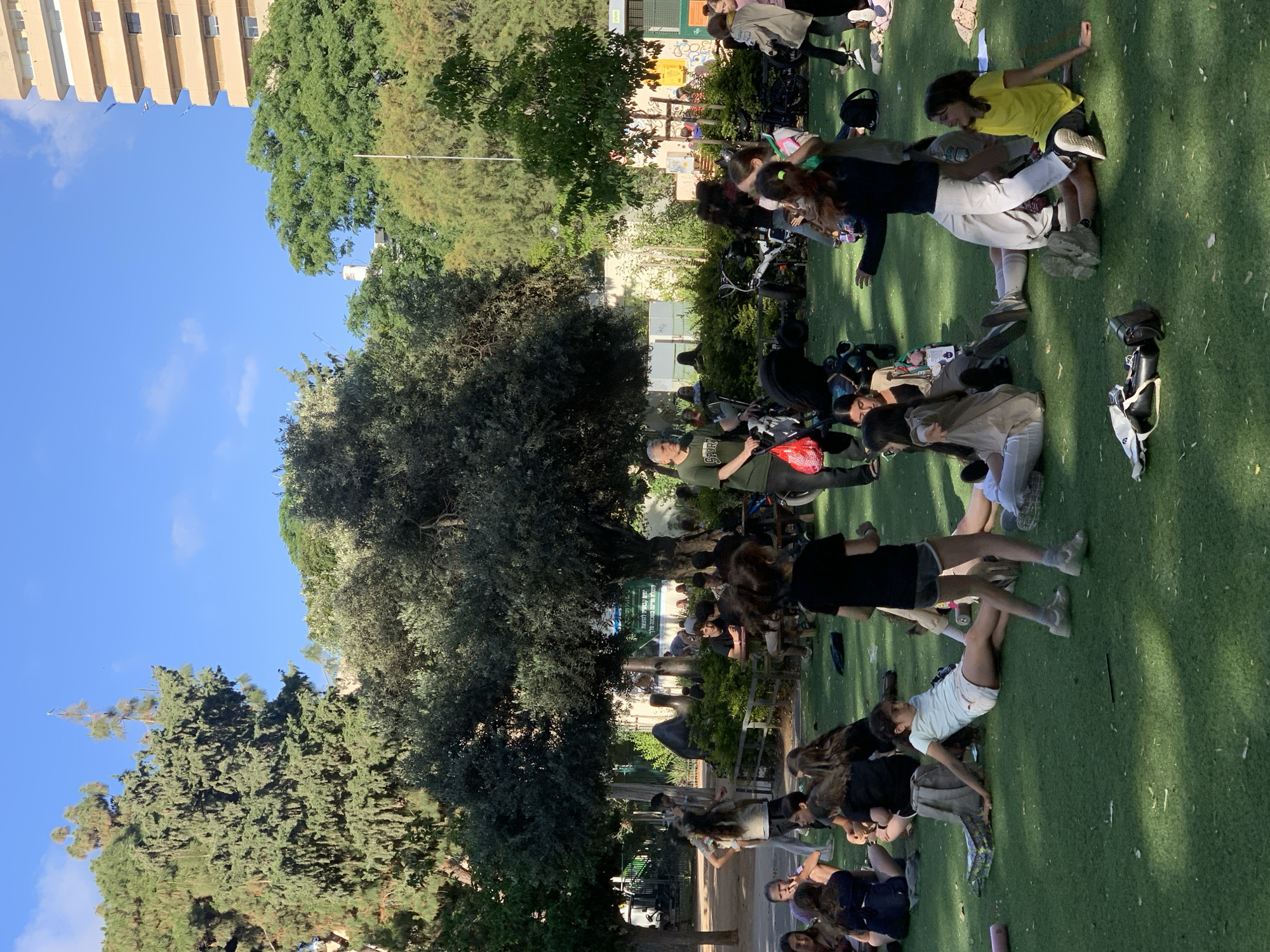 The image depicts a gathering of people in a park-like environment, likely in Tel-Aviv, Israel. A group of individuals, mostly young adults and children, are scattered on a green surface that could be artificial turf or closely cropped grass. Many are in various states of repose, lying down or sitting. A man in a green shirt and hat appears to be overseeing or instructing the group. Several bicycles and scooters are parked near the group, suggesting they arrived via personal transport.
Behind the group, there is a fence and some foliage, indicating a boundary to the park. Further back, a building is visible, suggesting an urban setting. The weather appears to be sunny, as evidenced by the clear blue sky and the bright light. The overall scene suggests an outdoor activity or gathering.