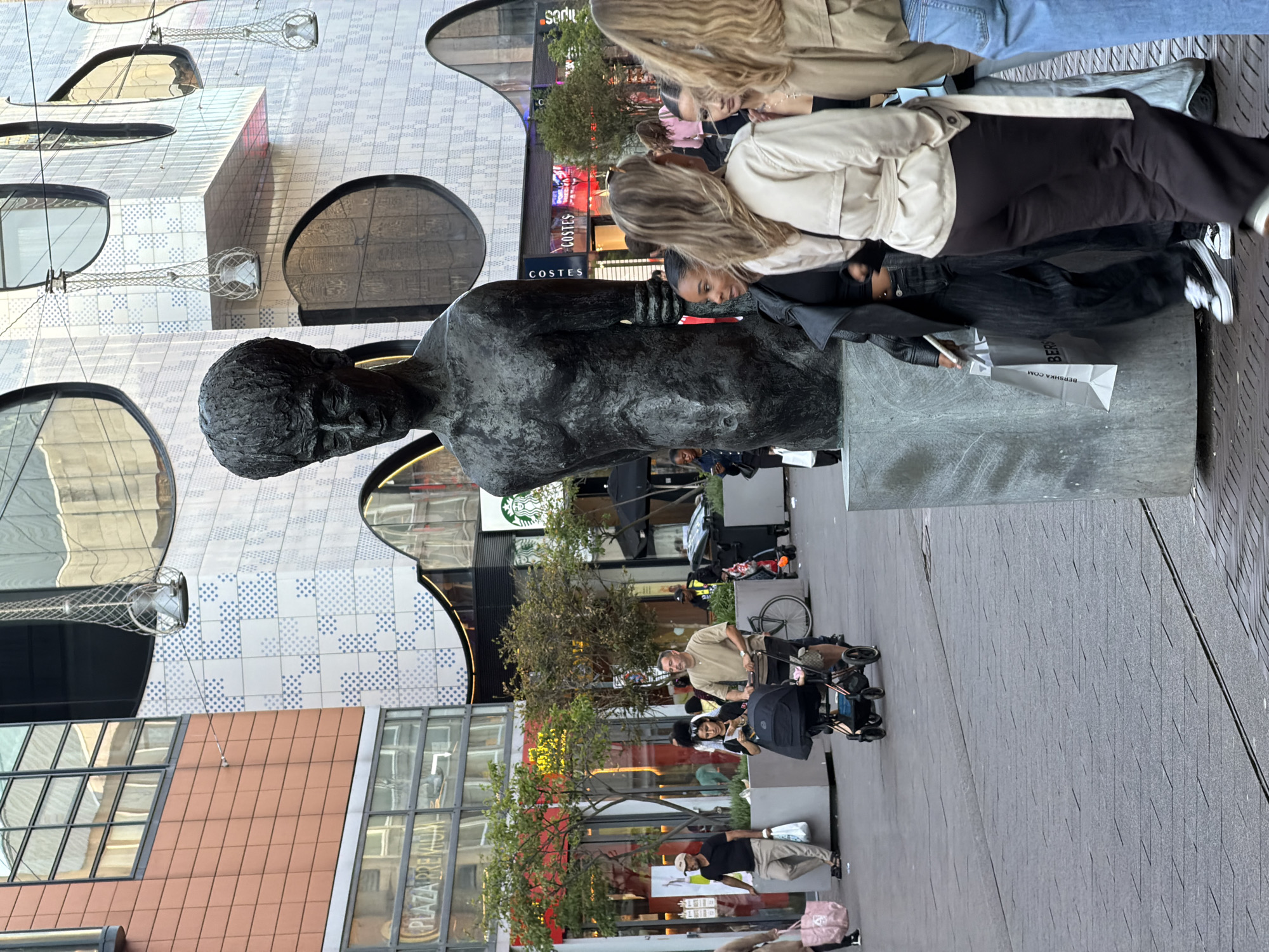 A full-body, medium-shot photograph shows a dark bronze statue of a man in the foreground, positioned in an urban plaza. The statue is of a nude male figure with a large head and wrinkled brow, standing on a grey, concrete base. Behind the statue, the plaza is paved with grey and black square tiles. In the background, a modern building with a white, patterned facade and large, dark circular windows dominates the scene. To the left of the statue, several people are walking on the plaza. A man in a tan shirt pushes a stroller with a child, accompanied by a woman in a black top. Further to the left, a man in a black t-shirt and light-colored pants walks with a shopping bag. Several shops with signs and storefronts are visible in the mid-ground, including "PLAZA PREMIUM" and "COSTES." In the far background, more of the modern building and sky are visible. The overall lighting suggests it is daytime, possibly late afternoon. The location is identified as The Hague, Netherlands.