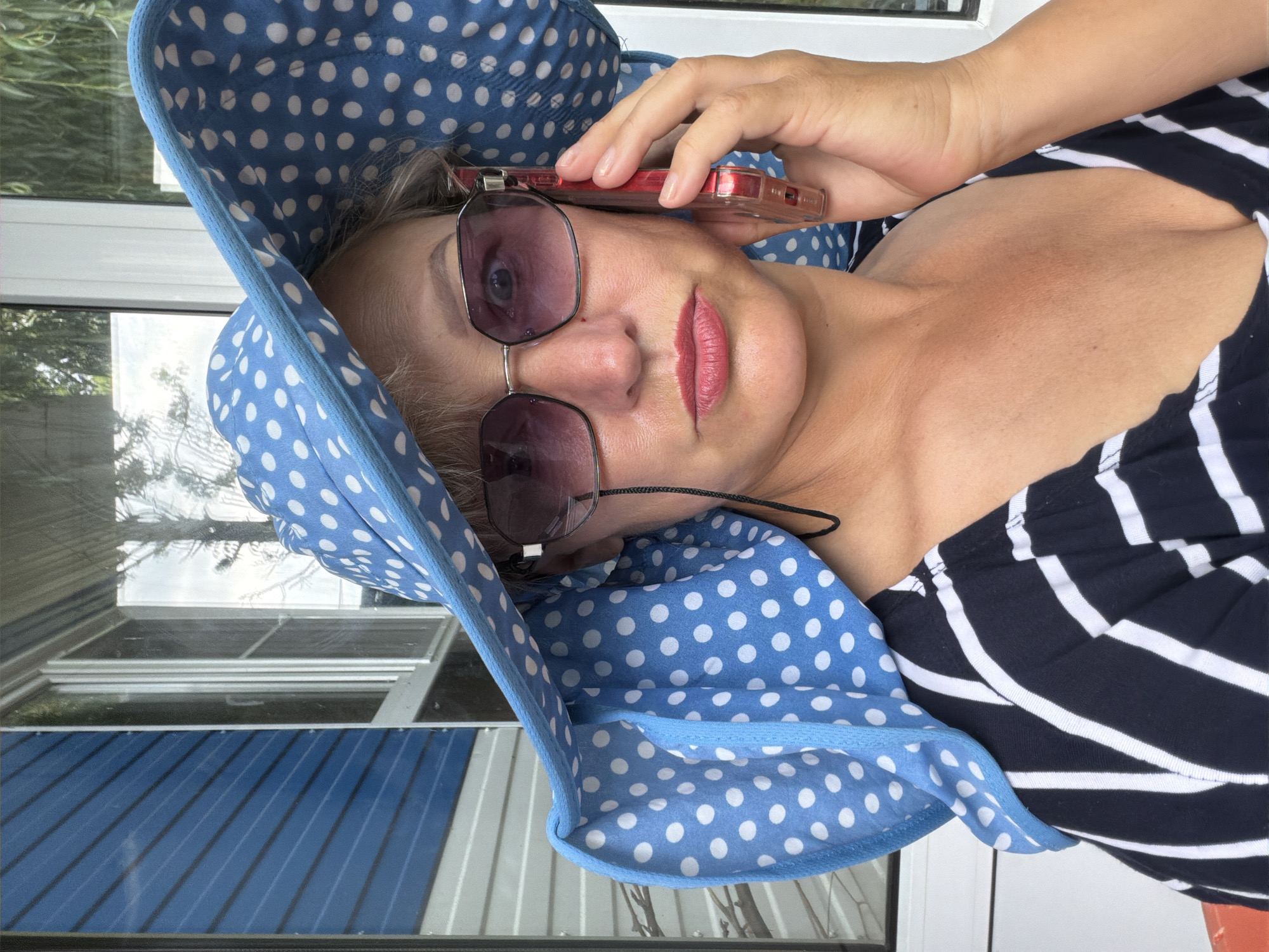 A woman is on the phone, wearing a large blue and white polka dot hat and a black and white striped shirt. She is outdoors, possibly on a porch or patio, with a blue-sided building and a window visible in the background. The woman is looking directly at the camera with a neutral expression. The scene appears to be in the city of Shepsi, Russia.