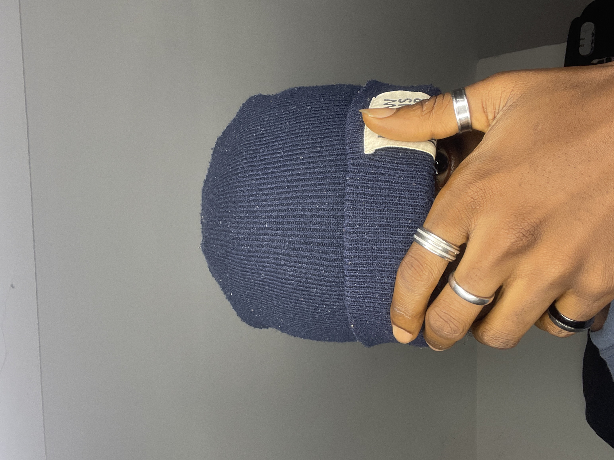 A close-up shot focuses on a person's hands and a dark blue knitted beanie. The beanie is worn on the head, obscuring most of the face, with only a glimpse of an eye visible through a gap. The beanie has a small, light-colored tag attached to it with some visible text. The person is wearing several rings on their fingers. On the thumb, there is a wide silver ring with a grooved design. The index finger has a thinner, smooth silver ring, and the middle finger sports a similar, slightly thicker silver band. The ring finger has a black band with a silver inlay. The skin tone is dark. The background is a plain, light gray wall, with some darker shadows to the right. The lighting appears to be indoor. The image is oriented sideways. There is no indication of the time of day or weather. No specific location cues beyond the general indoor setting are present.
