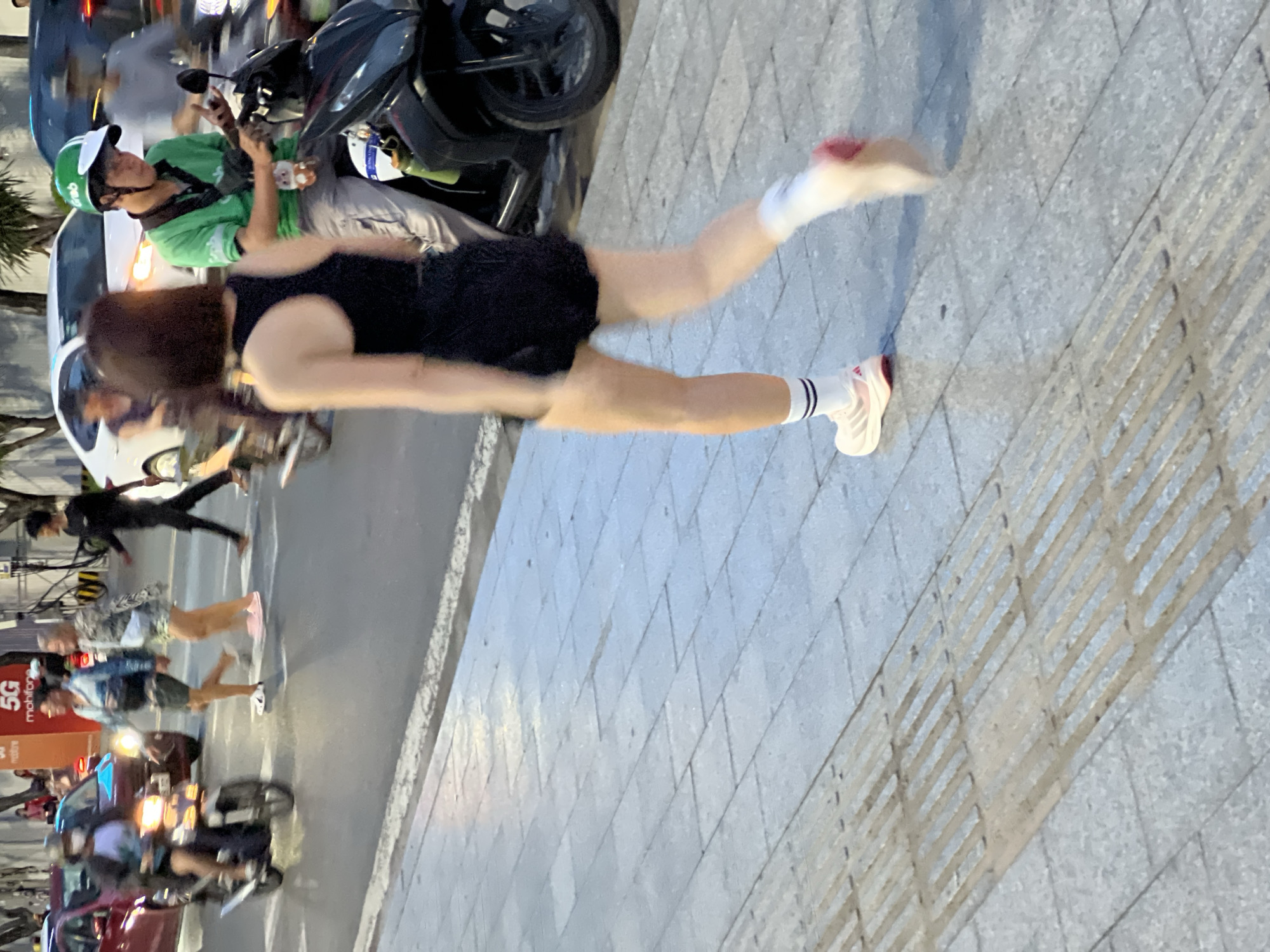 The image depicts a street scene in Nha Trang, Vietnam, focusing on a woman walking on a sidewalk. She is wearing a black tank top, black shorts, white socks with striped bands, and white and pink athletic shoes. The woman is in motion, with her legs blurred due to movement. Behind her, a man in a green shirt and helmet is riding a black motorcycle, looking down at his phone. Other people and vehicles, including cars and motorcycles, are visible in the background, suggesting a busy urban environment. The sidewalk is paved with grey tiles and has tactile paving for visually impaired pedestrians. The scene appears to be captured during daylight.