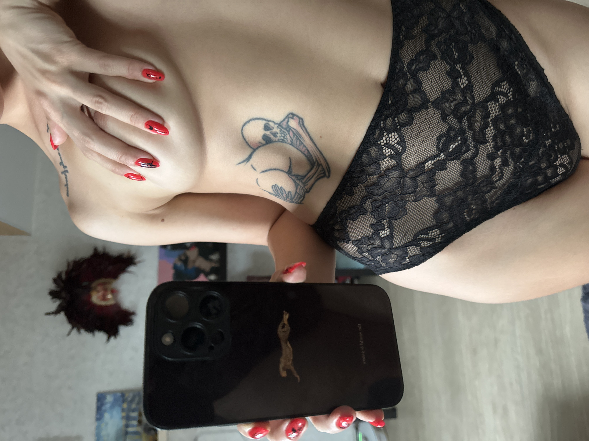 The media file shows a close-up view of a person's torso, focusing on their chest and abdomen. The individual has red painted nails and a tattoo on their side that appears to be a skull interacting with a stylized posterior. They are wearing black lace underwear. In the foreground, a hand holds a black smartphone, which is angled to capture a reflection of the person and their surroundings. The reflection shows a wall with several pictures or art pieces hanging on it, and a decorative item that resembles a dark feather boa. The overall lighting suggests an indoor setting, possibly a room in Vladimir, Russia. The image is a self-portrait, indicated by the presence of the smartphone and its reflection. The composition is intimate and focuses on aesthetic details such as the tattoo, manicure, and lingerie. No specific time of day or weather conditions are discernible. No other individuals are present in the frame.