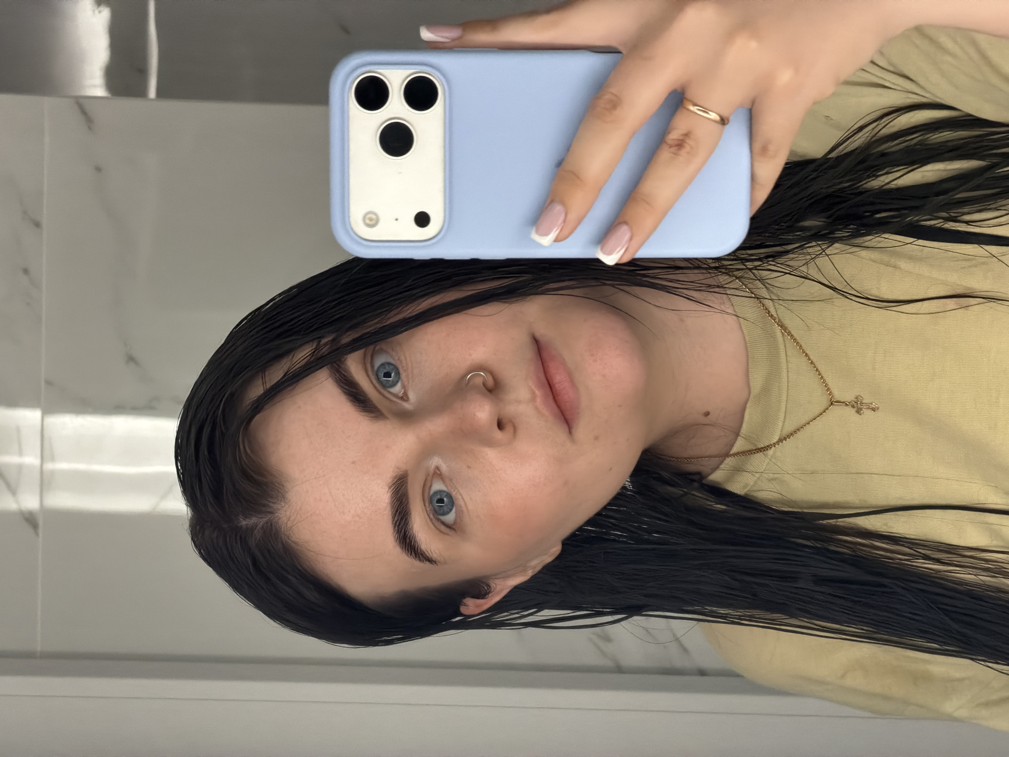 A selfie, potentially taken in a bathroom, features a fair-skinned woman with wet, dark hair. Her eyes are light blue. She has a nose ring. She is holding a light blue iPhone with three rear cameras, taking the photo. Her fingernails are painted with a French manicure. She is wearing a light yellow t-shirt and a gold necklace with a cross pendant. The background shows white and gray tiled walls. There's no verifiable evidence to place the location specifically in Chaikovsky, Russia, or any other specific city.