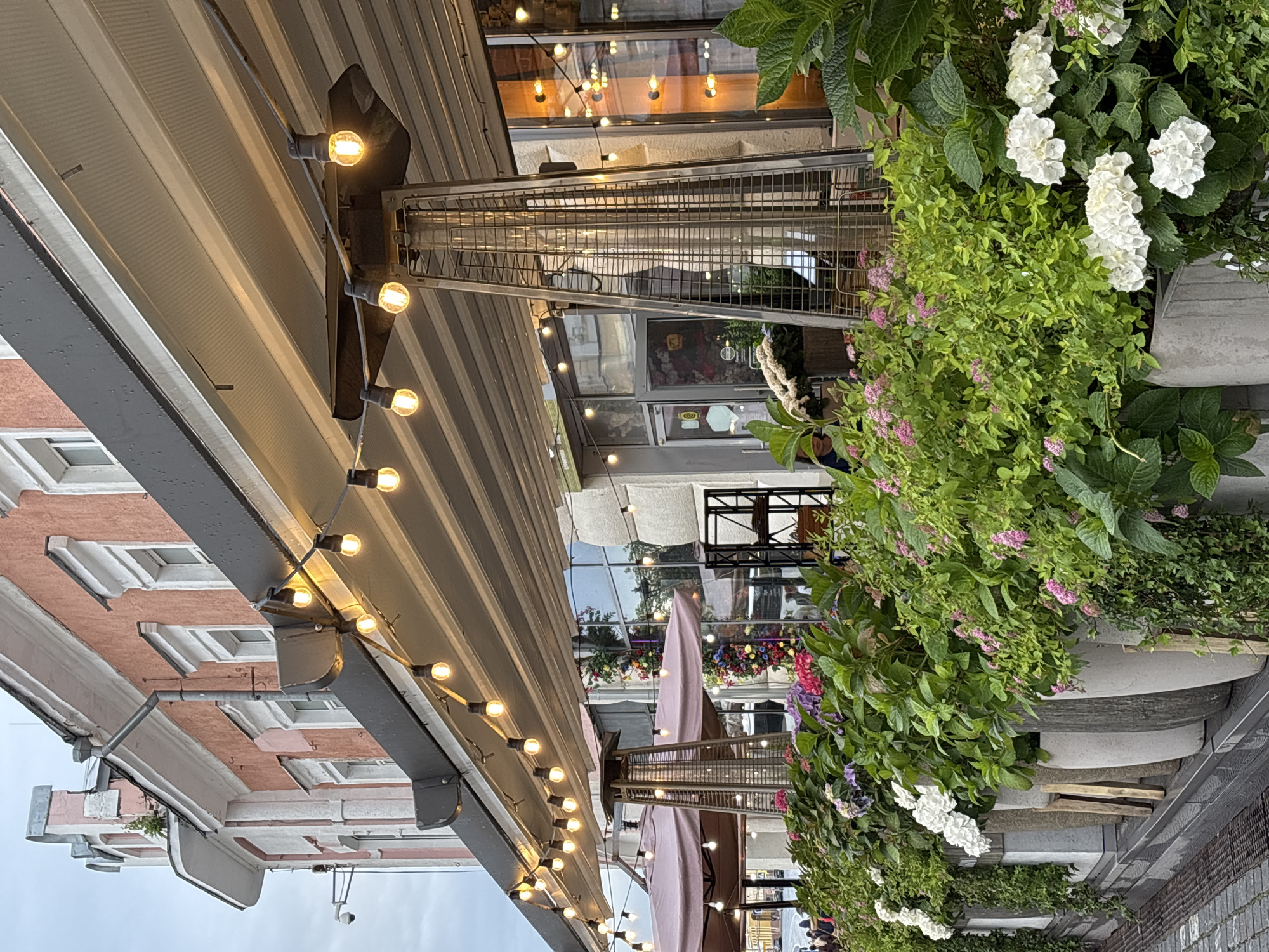 The image depicts the exterior of a cafe or restaurant in Nizhny Novgorod, Russia. The building has a salmon-pink facade with white trim around the windows. A dark awning extends from the building, supported by structural beams. Strands of Edison-style string lights are suspended beneath the awning, casting a warm glow. In front of the building, a row of large, grey planters overflows with lush green foliage and a variety of flowers, including white hydrangeas and smaller pink blossoms. A tall, pyramid-shaped patio heater stands between two pink patio umbrellas. Large glass windows and doors are visible behind the plants, reflecting the lights and greenery. The ground in front of the planters appears to be paved with dark cobblestones and a metal grate. A security camera is mounted on the side of the building. The sky is overcast.