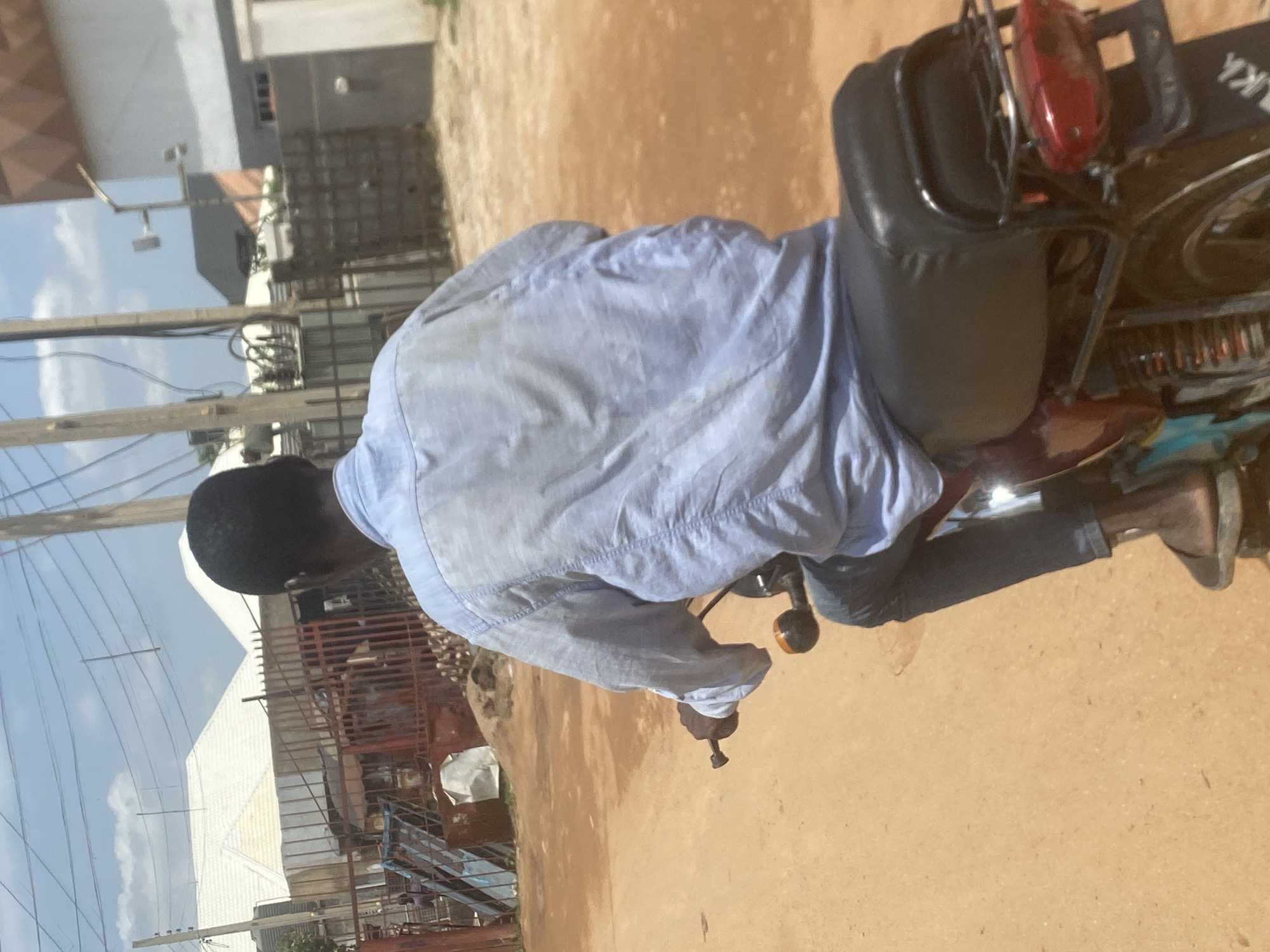 A person is riding a motorcycle on a dirt road in Lugbe, Nigeria. The person is wearing a light blue long-sleeved shirt and dark pants. The motorcycle is dark red with a black seat and a metal rack at the back. In the background, there are buildings, fences, and utility poles with wires. The sky is partly cloudy.