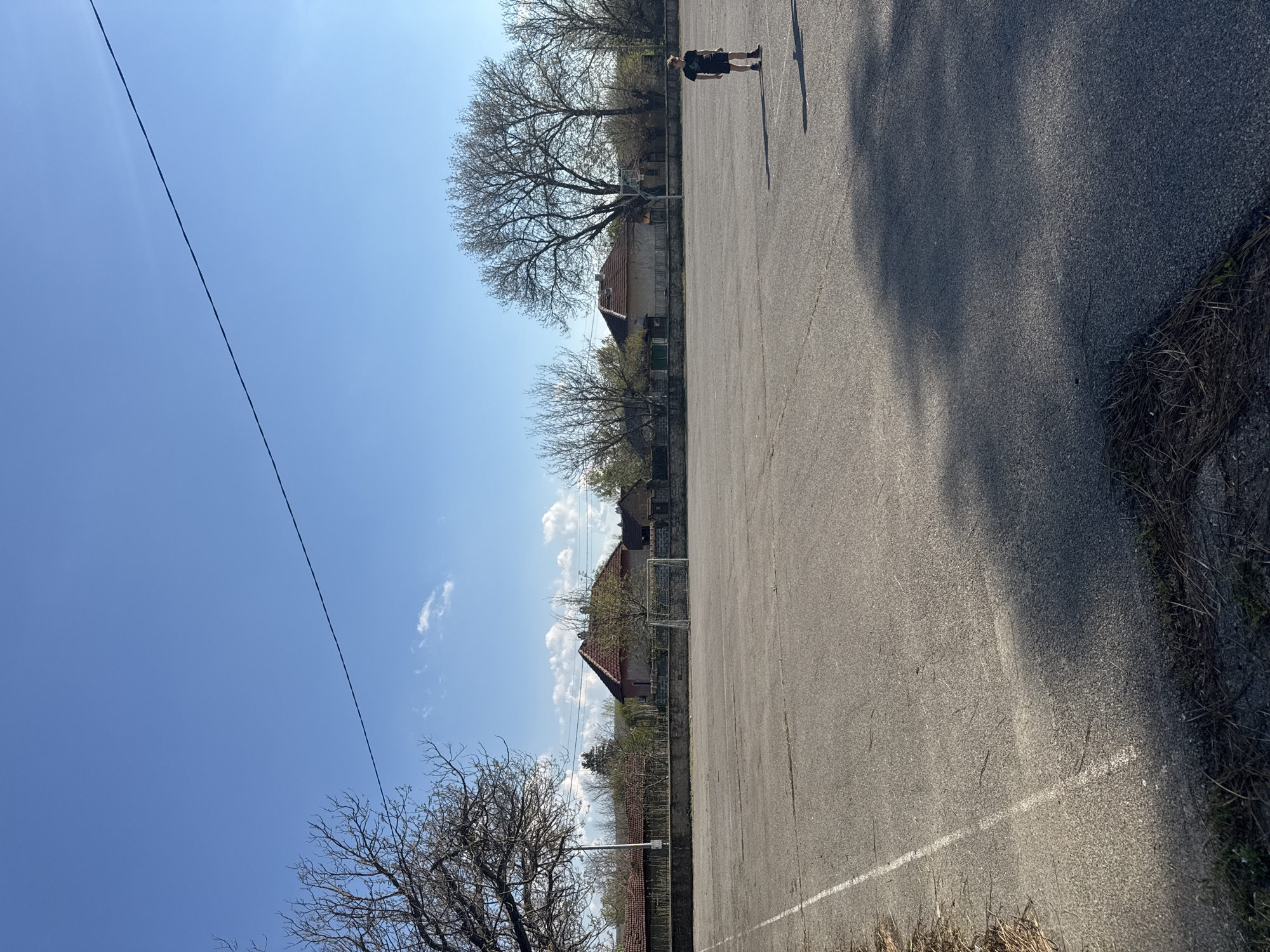 This image captures an outdoor scene, likely a community sports court or schoolyard in Lehchevo, Bulgaria, under a clear blue sky with scattered white clouds. The photograph appears to be rotated 90 degrees; when viewed in its intended landscape orientation, an asphalt court dominates the lower half of the frame.
On the right side of the court, a lone person, appearing to be a young male, stands facing left, wearing dark shorts and a dark t-shirt. His shadow stretches out behind him on the asphalt. A distinct white line is painted on the court surface, running horizontally across the lower part of the image. The ground texture is coarse asphalt, with areas of dried grass and soil visible along the edges.
The background features a row of bare deciduous trees, suggesting late autumn or early spring. Behind the trees, several modest houses with distinctive orange-tiled roofs are visible, contributing to a village or semi-urban setting. A white metal soccer or handball goal can be seen partially obscured by trees and a building in the mid-left background. Near the largest tree in the mid-ground, support for what appears to be a basketball hoop is also faintly visible, indicating a multi-purpose recreational area.
A power line or utility cable stretches diagonally across the upper left portion of the bright blue sky. The strong shadows cast by the person and potentially other objects imply bright sunshine, indicating it is daytime, likely mid-morning or afternoon. There is no discernible activity other than the person standing on the court. No visible text is present in the image.