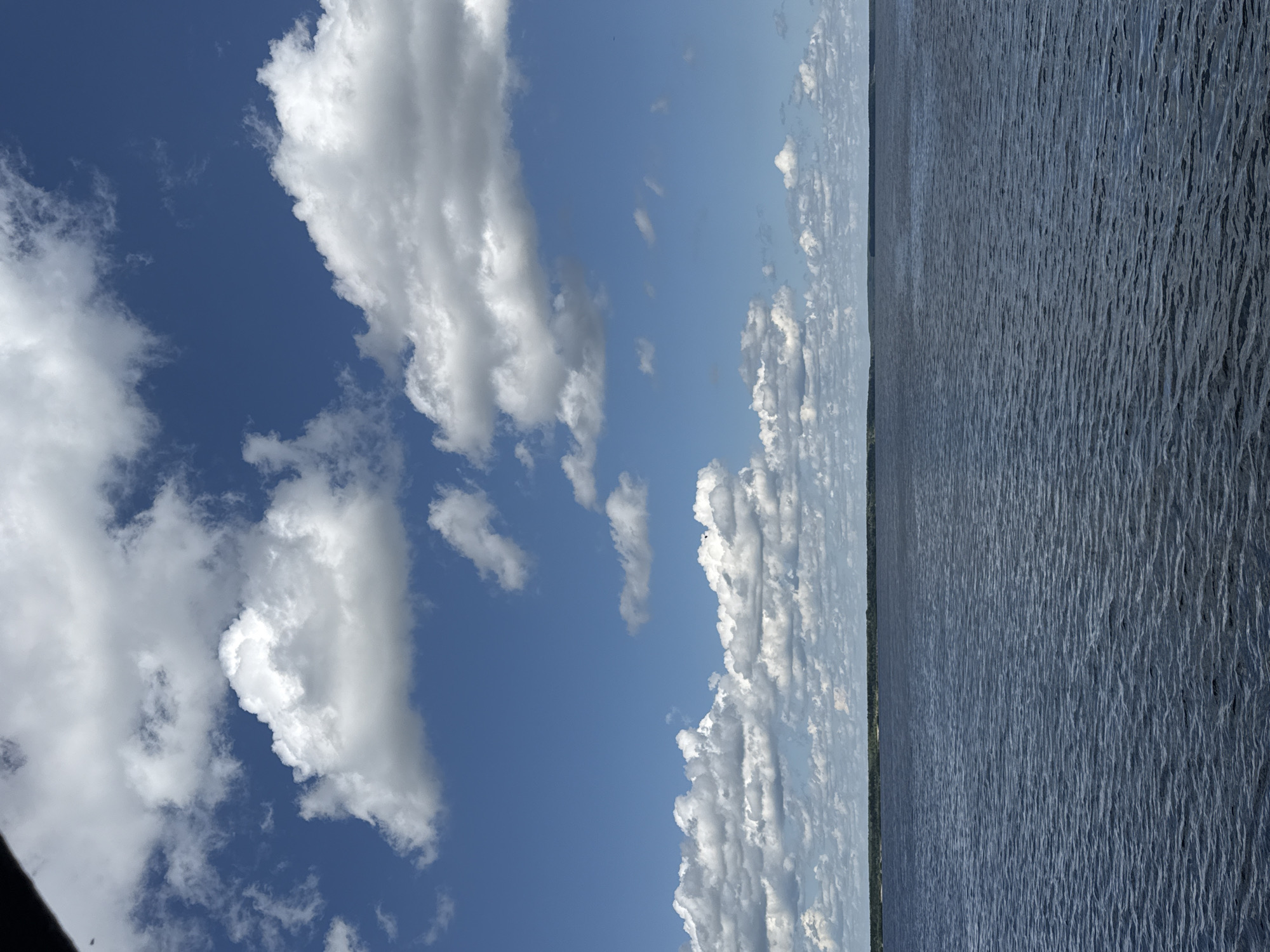 The image depicts a wide expanse of dark blue water under a bright blue sky filled with fluffy white clouds. The horizon is visible, showing a distant, tree-lined shore. The water's surface is rippled, indicating a gentle breeze. There are no people, animals, or man-made objects prominently featured in the scene. The overall impression is one of a calm, natural landscape, possibly a large lake or river. The location in Kozmodemyansk, Russia, is not visually identifiable from the image itself.