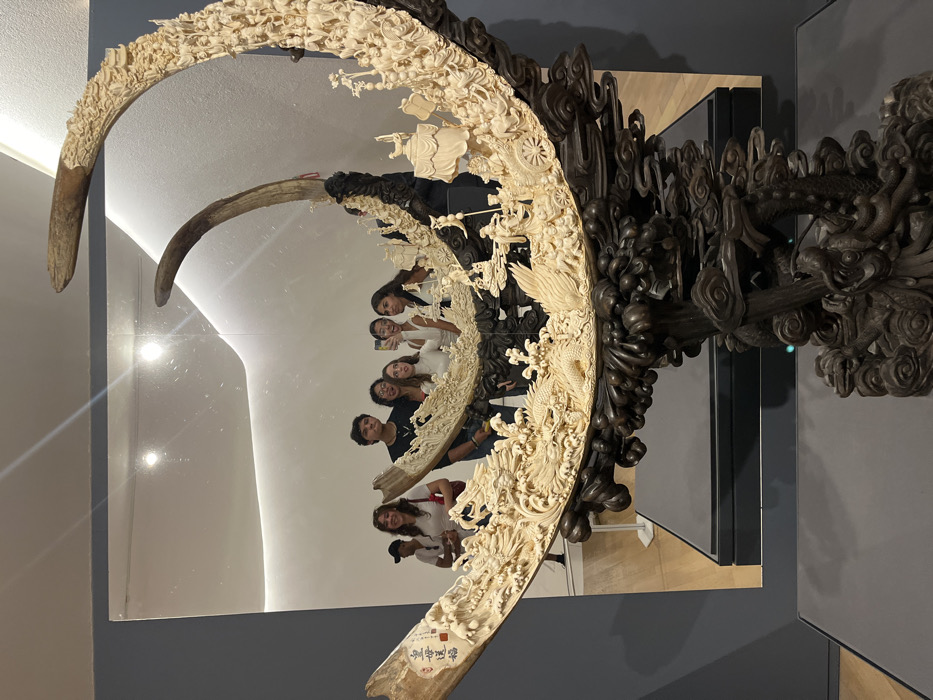 A large, ornately carved tusk-like sculpture is displayed in front of a mirror. The sculpture features intricate designs and figurines, and the mirror reflects a group of six people. The sculpture appears to be made of a light-colored material, possibly ivory, with a dark wooden support structure featuring dragons and other decorative elements. The background consists of a light-colored wall and a grey display panel, providing a gallery setting.