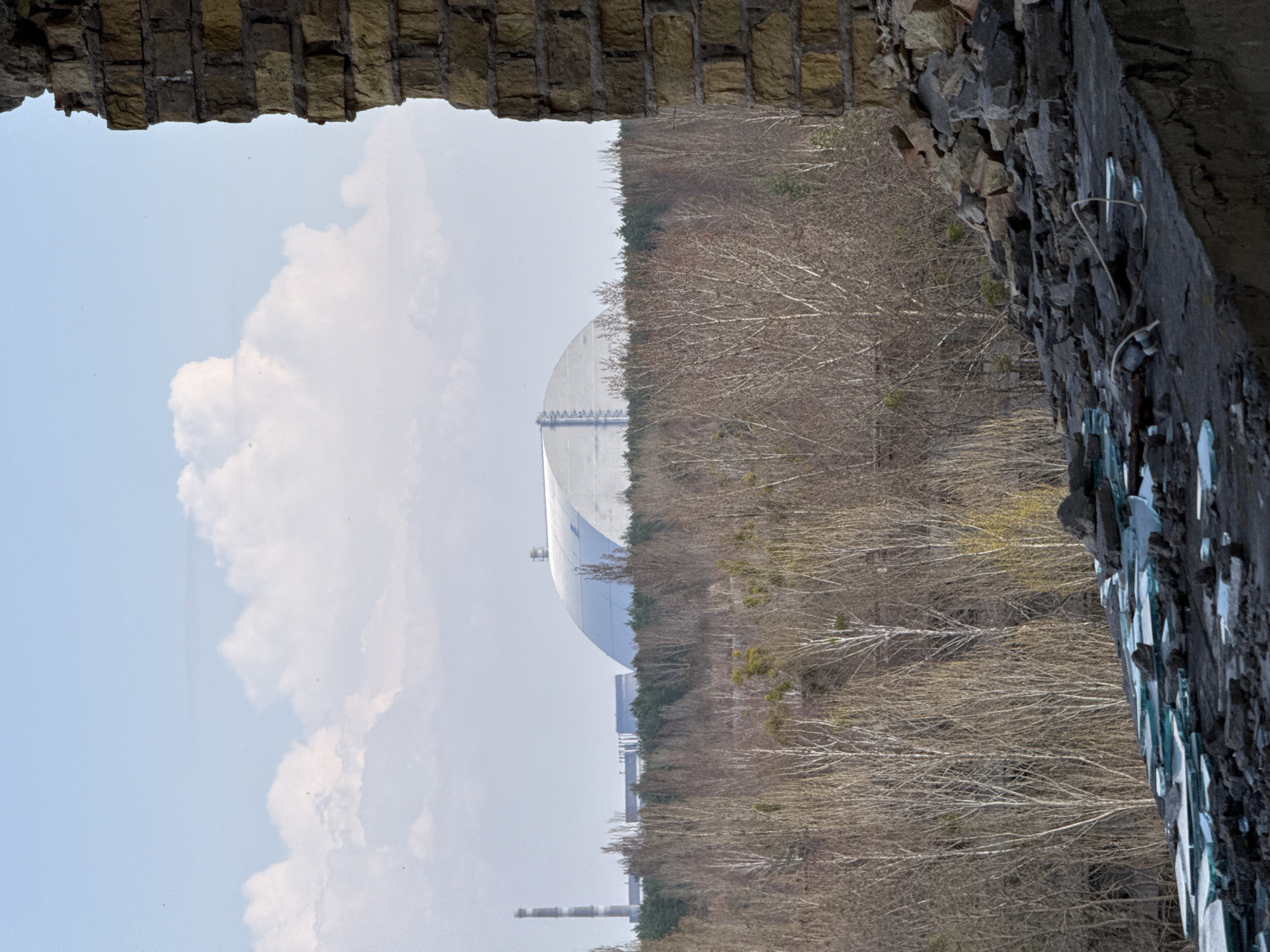 The image depicts the Chernobyl Nuclear Power Plant's New Safe Confinement (NSC), also known as the New Shelter or the Arch, viewed through a dilapidated opening in a structure, possibly a building or wall in Prypiat, Ukraine. The NSC, a large metal arch-shaped structure, dominates the central background. The sky is pale blue with scattered white clouds. The foreground features the edges of the crumbling structure, made of weathered brick and concrete. Trees without leaves are seen in the landscape between the NSC and the foreground structure, contributing to the bleak setting. There are no visible people in the image. The location is confirmed by the presence of the NSC, a defining structure of the Chernobyl Exclusion Zone near Prypiat.