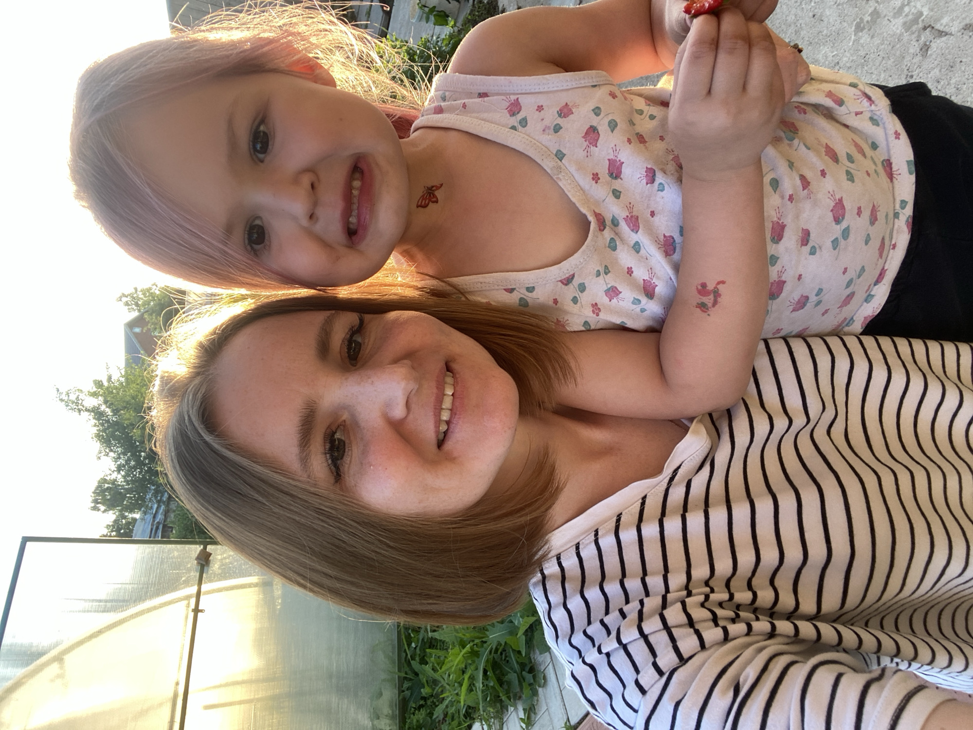 An outdoor, eye-level, medium-shot selfie captures a woman and a young child posed closely together. The woman, with short, auburn hair and freckles, smiles warmly at the camera. She wears a black and white striped t-shirt. To her right, the child, with light pink-toned hair, grimaces with a distressed expression. The child wears a light pink tank top adorned with a floral pattern and a temporary butterfly tattoo on their neck. Another temporary tattoo of a bird is visible on the child's left arm. The child is holding a red strawberry in their right hand. The background is illuminated by the warm glow of the setting sun, casting a bright light on the scene. To the left, a greenhouse structure is visible, with lush green foliage and a hint of rural architecture in the distance, suggesting a location in or around Chaikovsky, Russia. The ground appears to be paved with concrete or cobblestones.