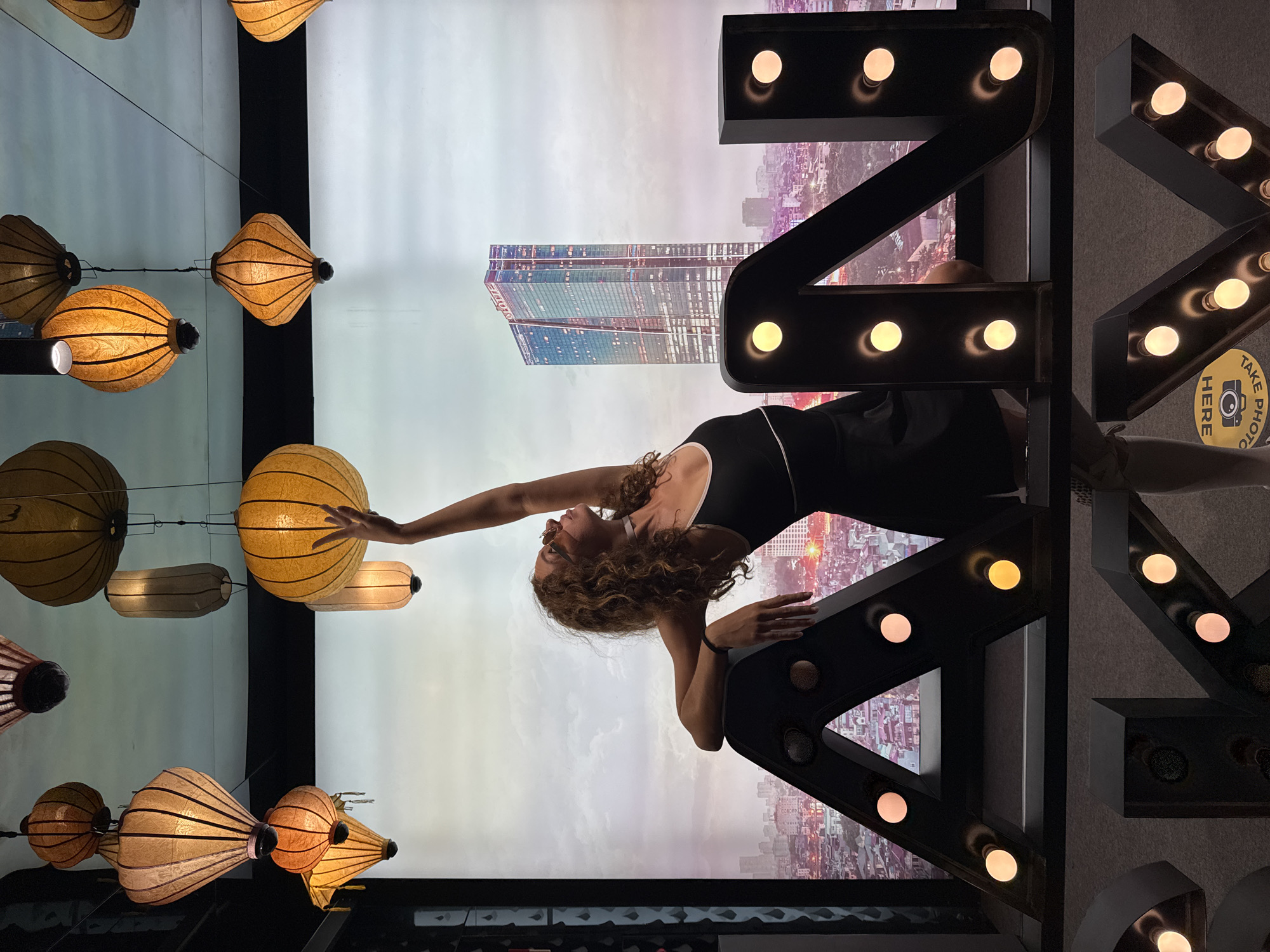 An adult female with curly brown hair, wearing a black sleeveless top and skirt, is posing in front of a backdrop depicting a city skyline, likely Hanoi, Vietnam. She is leaning on a large, black, illuminated letter "A" from a marquee sign. Her right arm is extended upwards, reaching towards a cluster of decorative paper lanterns hanging from the ceiling. The backdrop features a modern skyscraper in the distance, illuminated with numerous lights, set against a cloudy sky. Below the letter "A", partially visible, are other illuminated marquee letters forming part of a word. The overall scene appears to be an indoor installation or photo opportunity.