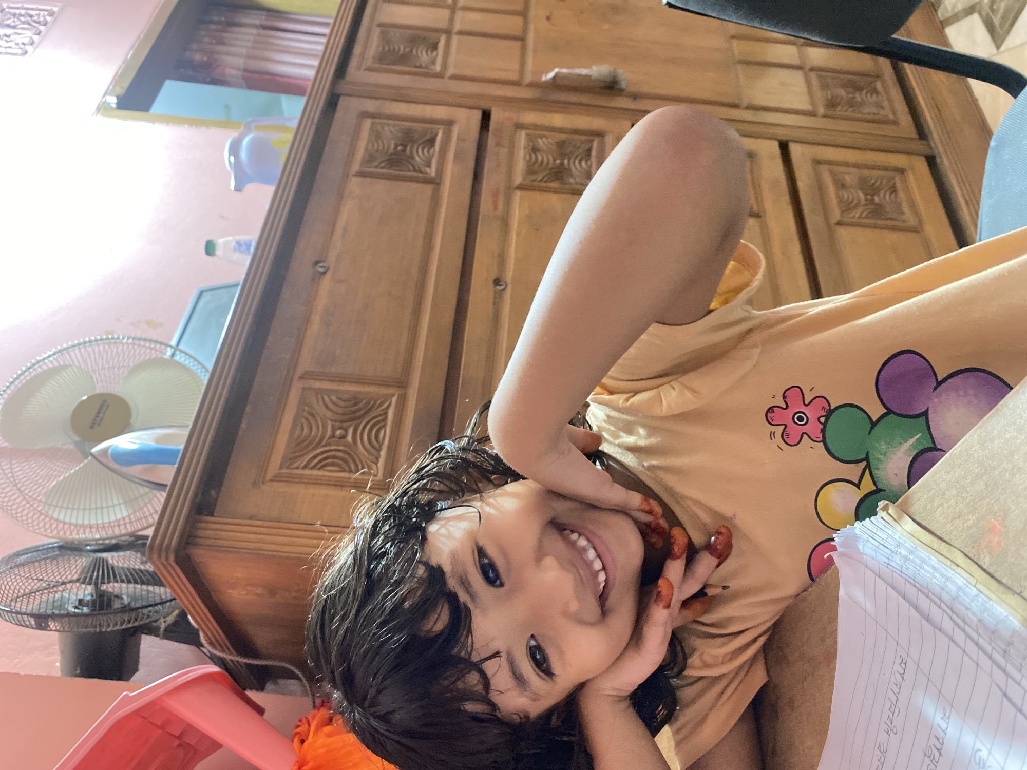 A young child, likely a girl, with dark, somewhat wet-looking hair, smiles broadly and directly at the camera. Her hands, adorned with intricate red henna (mehndi) patterns, are positioned playfully near her face. She is wearing a light yellow-orange t-shirt decorated with colorful cartoon flower designs in pink, green, and purple. Her expression conveys cheerfulness and happiness.
The scene is an indoor room, likely in a home in Dhaka, Bangladesh. In the background, a large, ornate wooden cabinet or wardrobe with carved panels stands prominently. To the left, two electric fans are visible: a larger white table fan and a smaller black fan below it, suggesting warm or hot weather conditions. The walls are painted a soft pinkish-peach color.
To the child's right, an open notebook with handwritten text is visible, indicating activities such as studying or writing. A partially legible phrase on the page reads "আলহামদুলিল্লাহ" (Alhamdulillah) in Bengali script. A small pencil is also seen. Other objects include a pink plastic stool or chair part in the bottom left, and partially visible containers and a water bottle on a shelf in the background. The photograph is taken from a low angle, looking up at the child, who appears to be resting on a low surface. The presence of henna on her hands suggests a special occasion or cultural celebration.