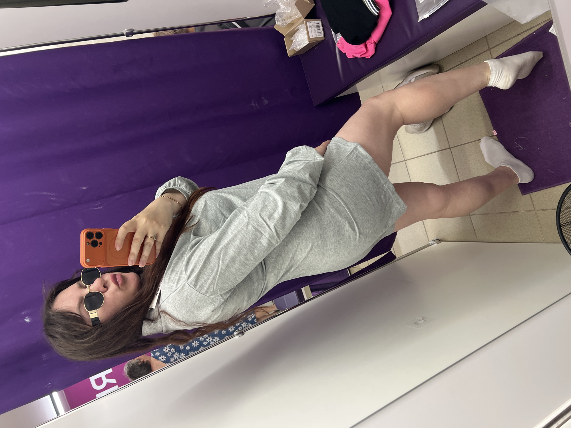 A woman is taking a selfie in a fitting room. She is wearing a light gray long-sleeved dress, white socks, and beige sneakers. She has on gold-rimmed sunglasses with black lenses. In the reflection of a mirror, another person can be seen wearing a blue shirt with a white floral pattern. The fitting room has a purple curtain in the background and a purple bench with some clothing on it. A white door is visible on the left side of the image. The flooring appears to be tile. The location is specified as Noviy, Russia, but this cannot be verified from the image itself.