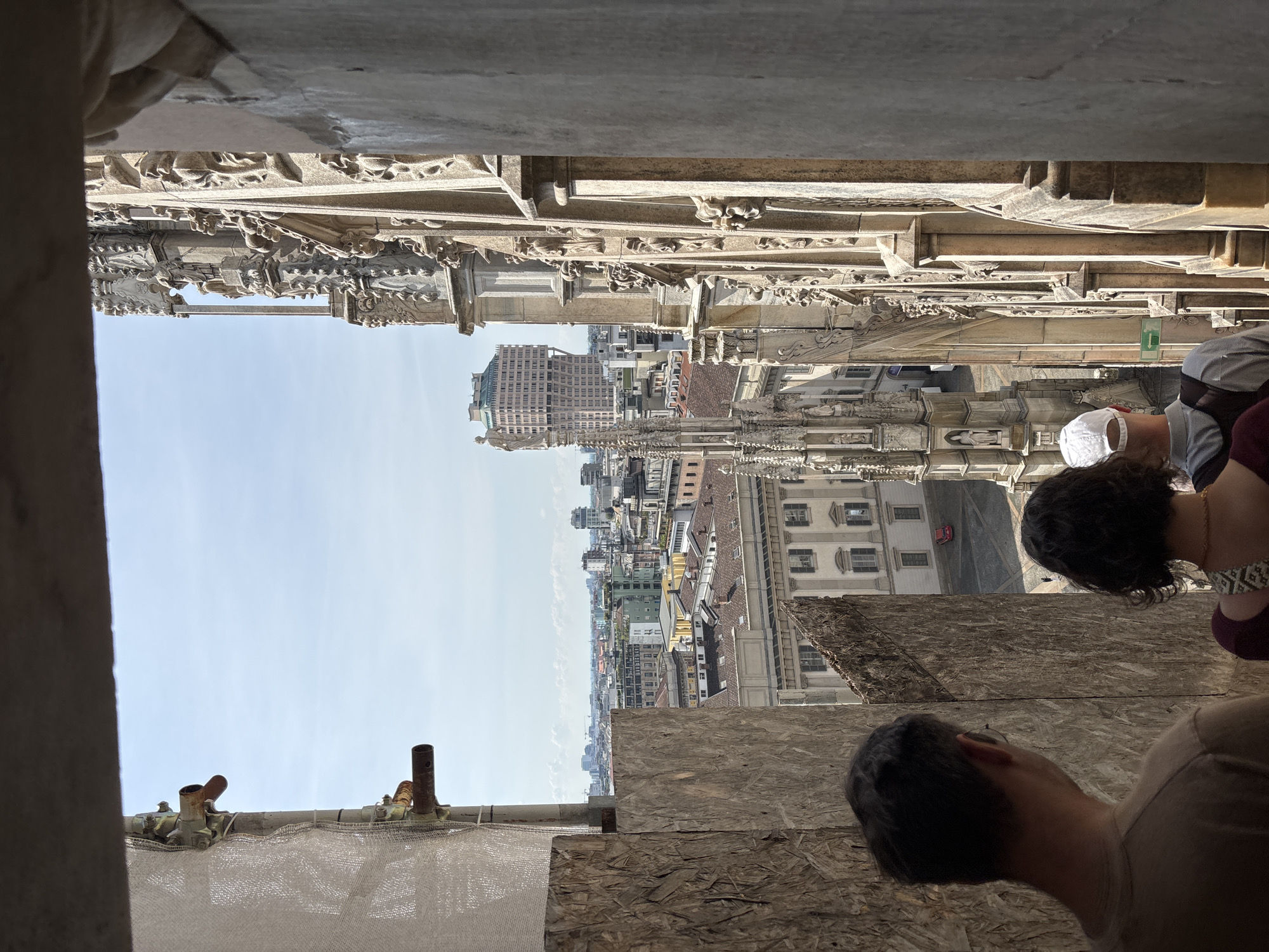 Here is a description of the image:
The image shows a view from the Duomo di Milano (Milan Cathedral) in Milan, Italy.
**Environment:** The scene is mostly outdoors, with a clear blue sky. The view extends over the city of Milan, showing a dense urban landscape with a mix of buildings, including a high-rise office or residential building in the distance. The cathedral's ornate stonework and architectural details are visible in the foreground, framing the cityscape. Temporary-looking plywood barricades can be seen on the floor, with some metal tubes visible.
**People:** At least three people are partially visible in the foreground of the image. Two are standing close together, possibly a couple, while a third person is partially visible to the left, wearing a white hat. They appear to be tourists or visitors looking out over the city.
**Objects:** Beyond the buildings, the Duomo features ornate stone carvings and gothic architectural details. Metal tubing runs along the floor, with some sort of white netting visible as well.
**Actions and Interactions:** The primary action appears to be the people looking out over the city from the vantage point of the cathedral. The framing of the image suggests they are tourists enjoying the view and taking in the scenery.