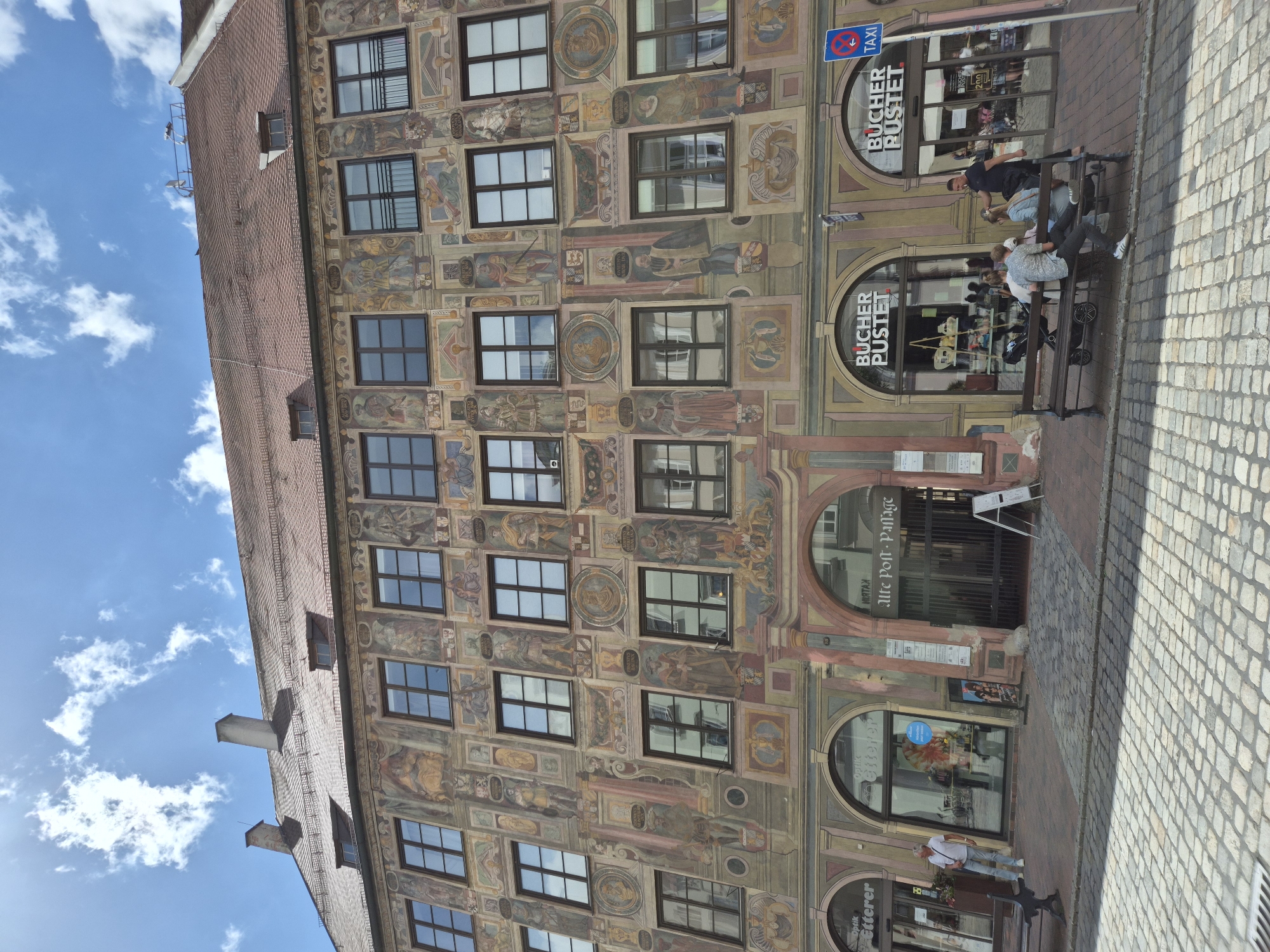 This image depicts the Renaissance-era Rathaus (Old Town Hall) building at Altstadt 26 in Landshut, Germany, on a clear day under a blue sky with scattered white clouds. The multi-story structure features a reddish-brown tiled gabled roof with two prominent chimneys and several smaller dormer windows. The entire facade is extensively decorated with faded, multi-colored frescoes or sgraffito, depicting numerous full-body figures, often in period attire, within architectural frames, alongside circular medallions featuring busts. Inscriptions, partially legible, are integrated into the design.
The building's upper floors are punctuated by numerous rectangular windows with dark frames, reflecting the sky and surrounding environment. The ground floor presents multiple arched entrances. On the left, two arched shopfronts belong to "Optik Etterer," displaying eyeglasses and promotional material. A man with gray hair, wearing a white short-sleeved shirt and light blue jeans, stands near the left shopfront. In the center, a large arched entryway with a dark metal gate leads to the "Alte Post-Passage," with the number "26" visible above the arch. To the right, two more arched shopfronts clearly display the name "Bucher Pustet," indicating a bookstore.
The foreground consists of a cobblestone street surface, primarily light gray and tan. Two dark-colored benches with wooden slats are positioned along the sidewalk. The left bench is unoccupied. The right bench is occupied by a group of five individuals. A man in a dark blue polo shirt stands behind the bench, interacting with those seated. A man in a patterned short-sleeved shirt and dark pants sits on the bench, along with three other individuals, one of whom has a baby stroller beside them. A blue circular sign with a red diagonal line, indicating "No Taxi" or "Taxi Stand," is visible on a pole to the right, near one of the "Bucher Pustet" entrances. Shadows cast by the building and individuals indicate strong sunlight from the right side of the frame.