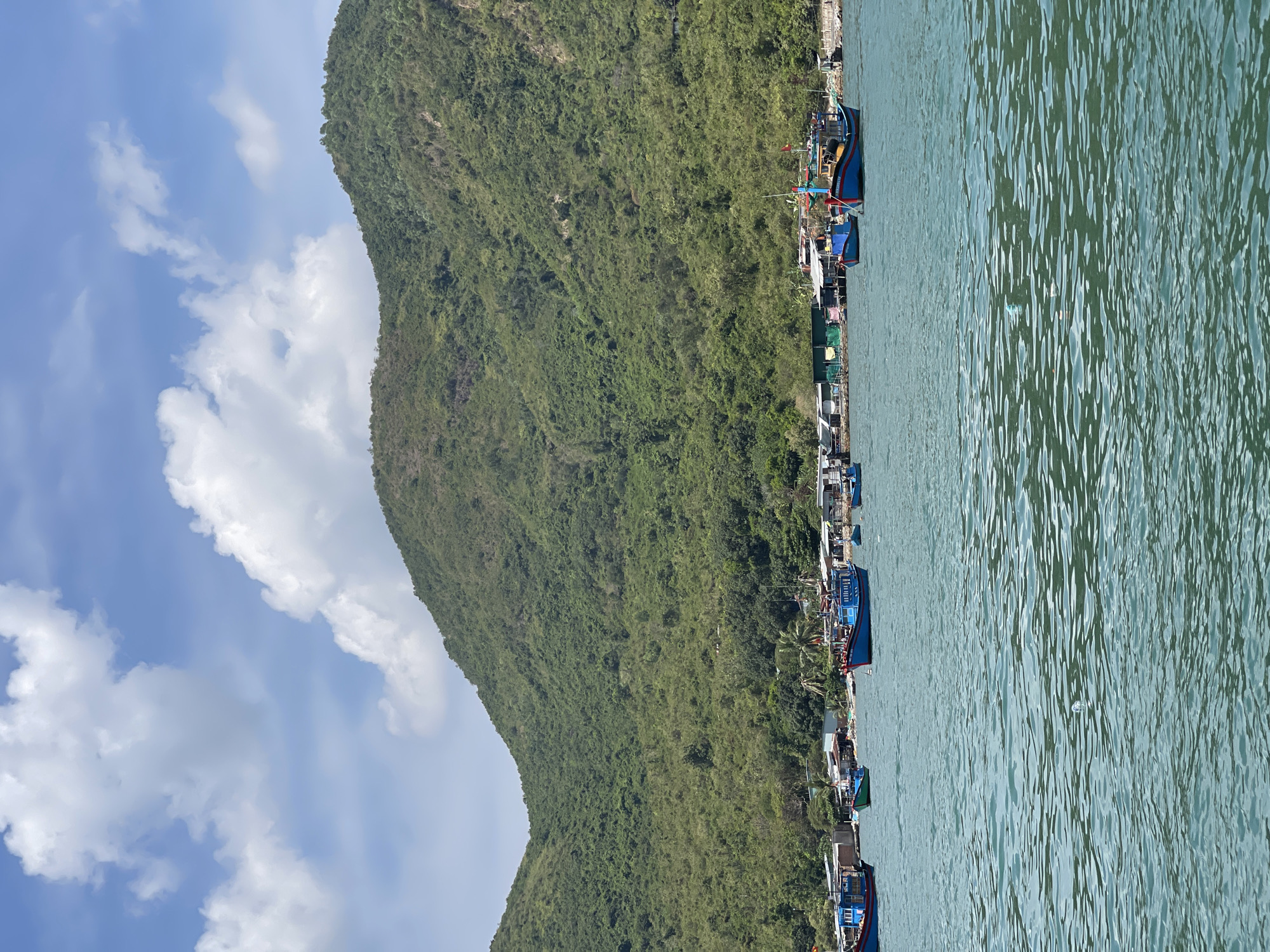 The photograph depicts a coastal scene with elements of nature and human activity. A large, lush, green mountain dominates the left side of the image. Along the base of the mountain, a small settlement is visible, interspersed with trees and featuring several buildings. A line of boats, mostly blue, are docked along the shoreline, suggesting a fishing community. The right side of the image is filled with water, reflecting the sunlight and the sky. The sky itself is partially covered with white clouds against a blue backdrop. The image appears to be taken from the water, creating a perspective that emphasizes the scale of the mountain and the proximity of the settlement to the sea. The location is consistent with the coastal topography of Nam Nha Trang Ward, Vietnam.