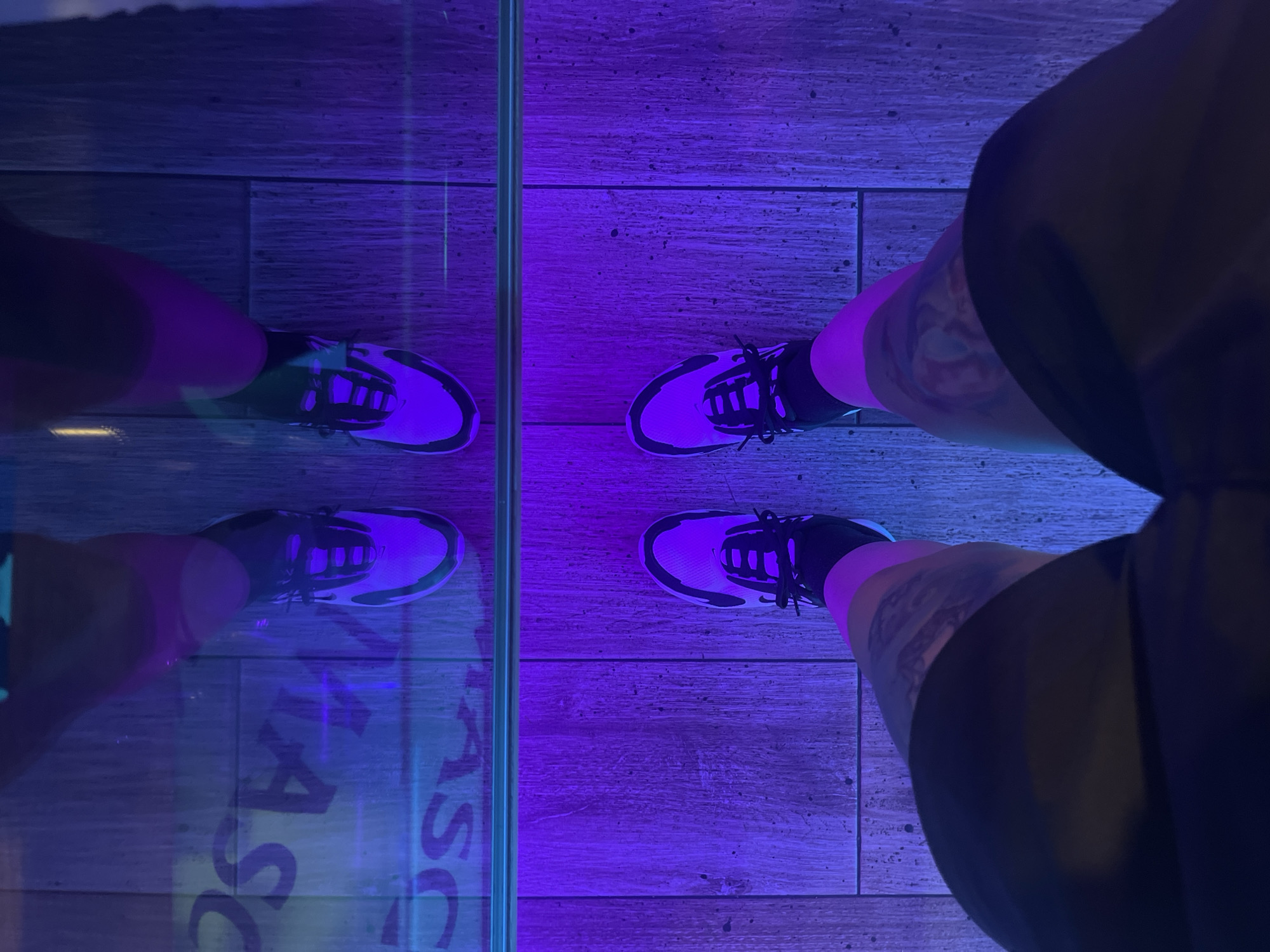 This image shows a person's legs and feet, wearing athletic shoes and dark shorts. The scene is set on a wooden floor that is illuminated by purple and blue lights. A clear vertical barrier, likely glass, divides the frame, reflecting the person's legs and feet. The reflection reveals a faint, stylized text on the floor beneath the glass barrier. The overall mood is modern and possibly athletic, with the lighting creating a dynamic and somewhat abstract atmosphere. The location context of Barcelona, Spain, doesn't offer specific visual cues in this image. There are tattoos visible on the person's legs.