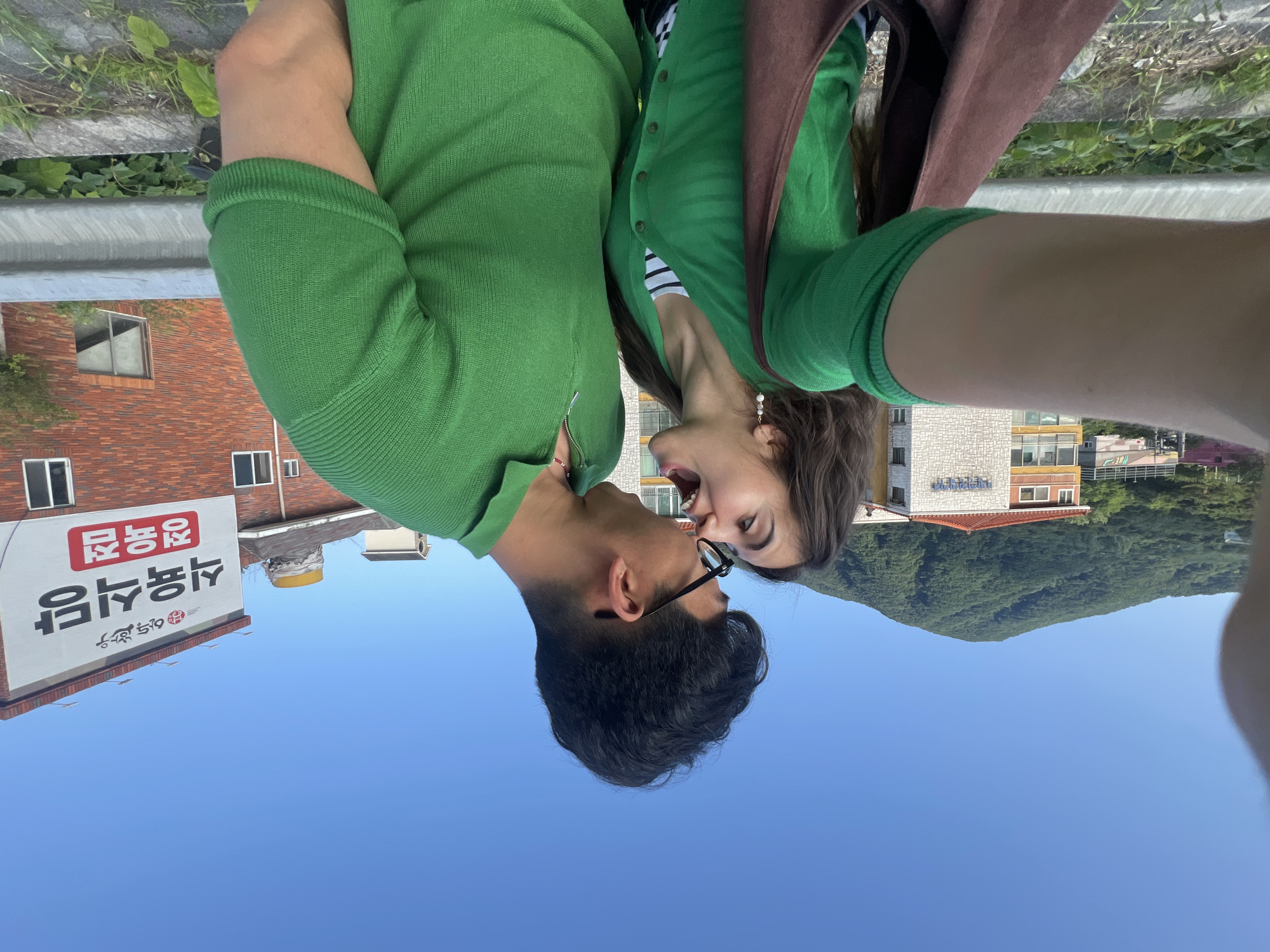 A couple is taking a selfie outdoors, with a man leaning in to kiss a woman. They are both wearing green tops. In the background, there is a sign with Korean writing, a brick building, a guardrail, greenery, a mountain covered in trees, and a clear blue sky. The image appears to be taken in Dongmyeong-myeon, South Korea.