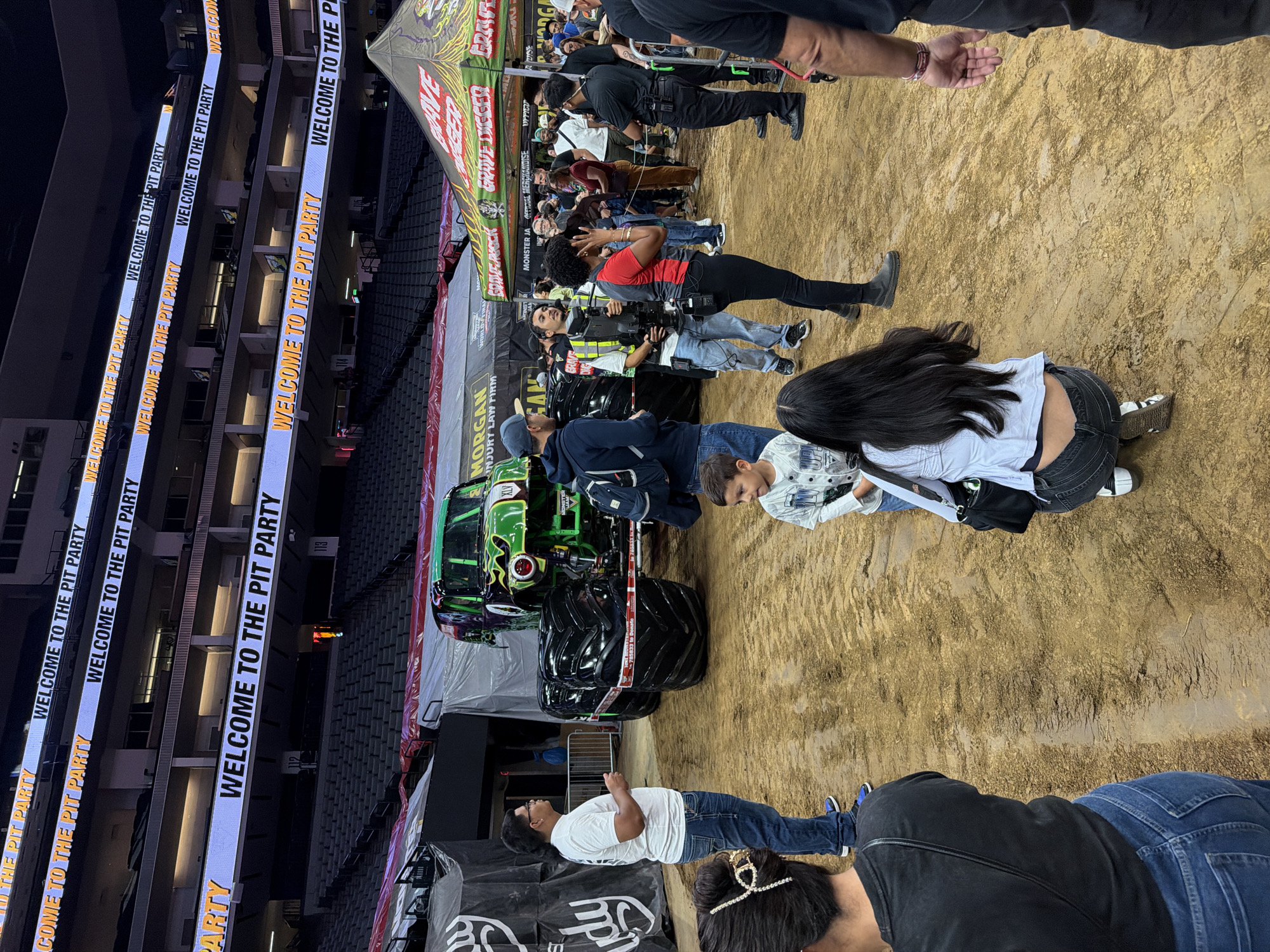 This image depicts a monster truck event at a large indoor venue, likely an arena in Sacramento, California. The foreground shows several people milling around on a dirt surface, with tire tracks visible. A green and black monster truck is prominently positioned in the middle ground. In the background, stadium seating is visible, and large digital billboards display the text "WELCOME TO THE PIT PARTY." A green tent with "GRAVE DIGGER" branding is also present, suggesting a Grave Digger themed event. The overall atmosphere appears to be that of a public gathering focused on monster trucks, likely part of a larger event or competition.