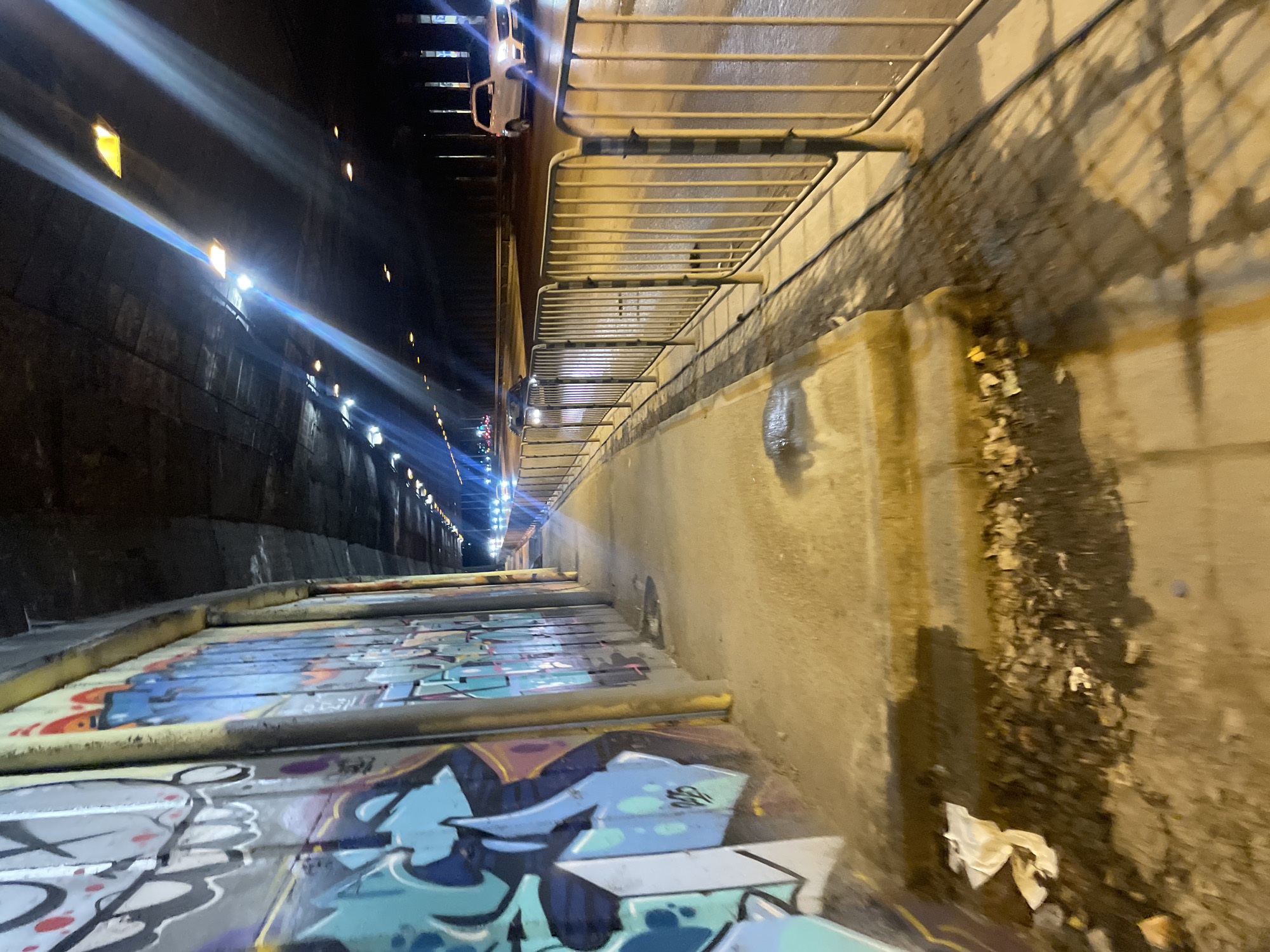 Captured from a low angle, the photograph depicts a section of the underpass beneath Sofia University in Sofia, Bulgaria. The right side of the image features a worn, concrete wall with water stains and litter. A metal railing runs along the top of the wall. To the left of the wall is a walkway covered in graffiti, with metal bars delineating the edge. Beyond the walkway, the tunnel recedes into the distance, illuminated by intermittent lights. A car is visible, parked on the right near the top of the frame.