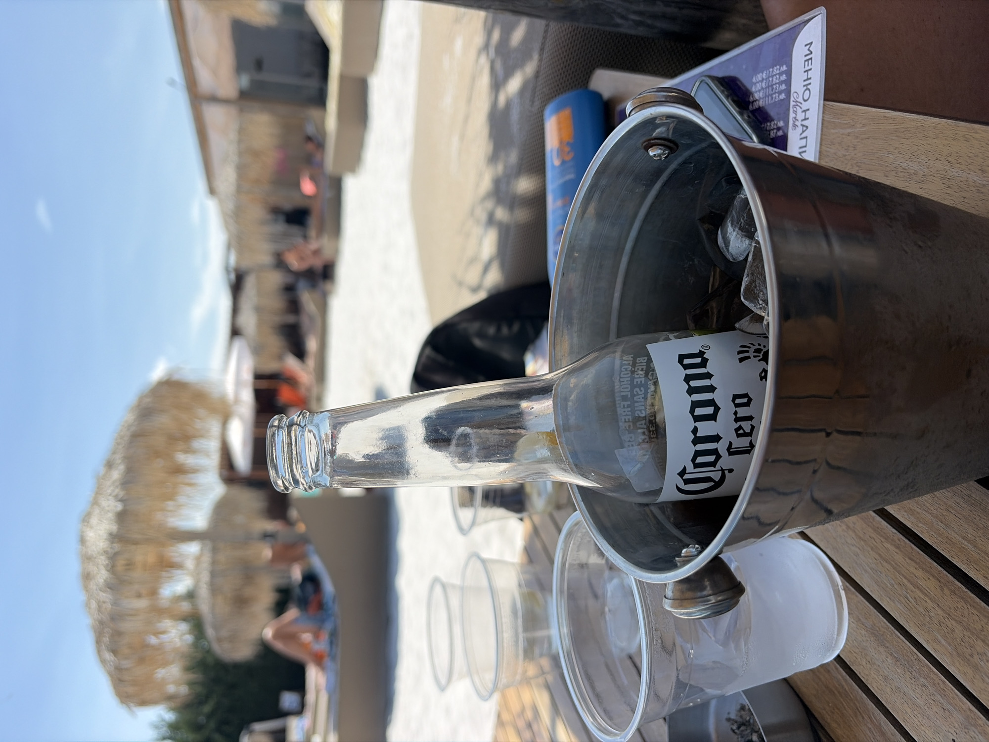 A high-angle medium shot captures a close-up of a silver ice bucket on a wooden table. Inside the bucket, a clear glass bottle of Corona Cero beer is partially submerged in ice. To the left of the bucket, there are three clear plastic cups, two of which contain a clear liquid and ice. The background is out of focus, but suggests a beach or poolside resort setting with thatched umbrellas, lounge chairs, and people relaxing. The sky is a clear, bright blue. The scene is set outdoors on a sunny day. The location is identified as Kazichene, Bulgaria.