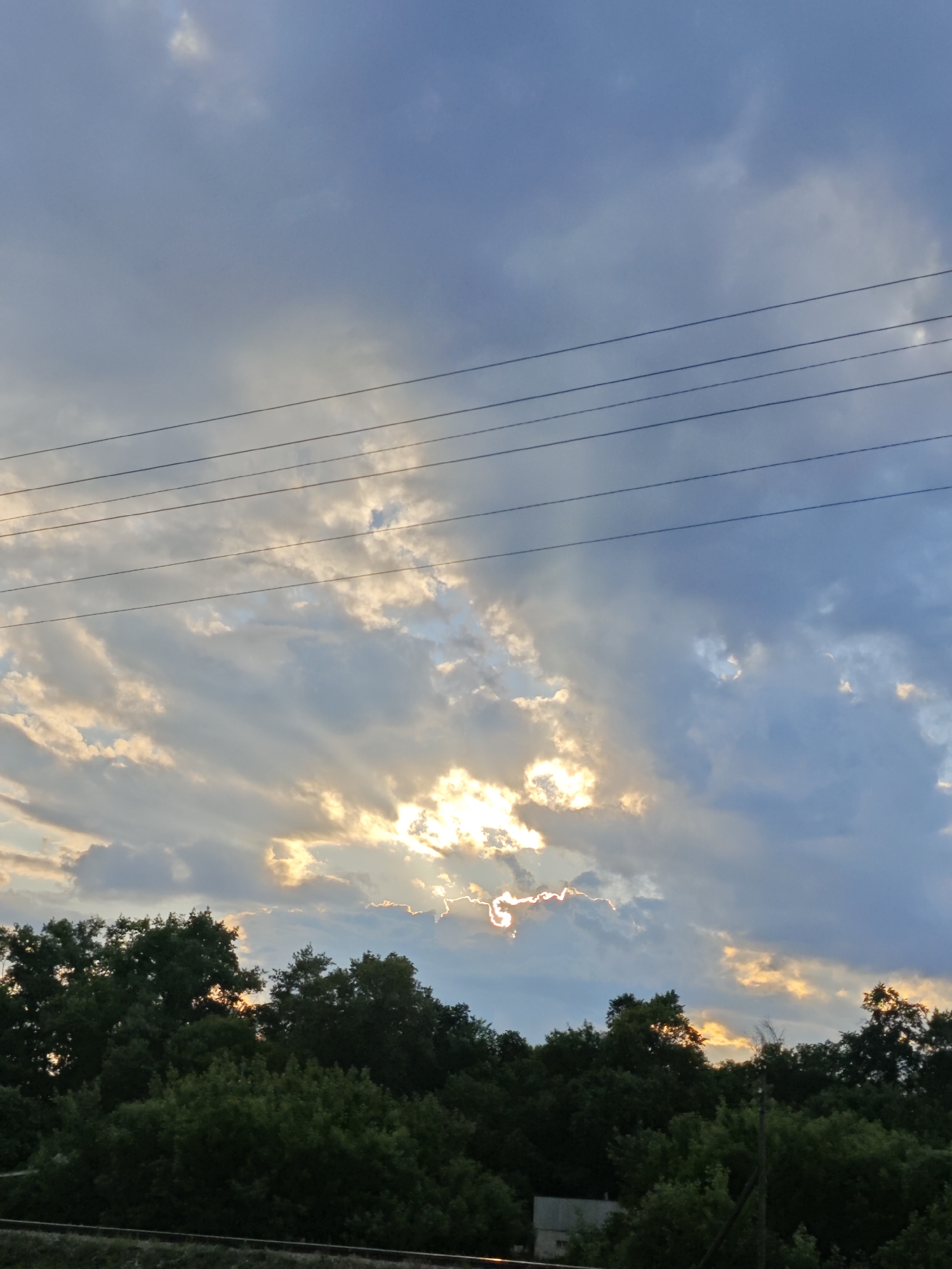 The image depicts a sky with dramatic clouds, illuminated by the sun breaking through. Several power lines are visible in the upper portion of the frame. Below the sky is a dense line of green trees. In the lower right corner, a utility pole is partially visible, and a small, light-colored building can be seen behind the trees. A railway track runs along the bottom edge of the image. The scene appears to be an outdoor, rural or semi-rural setting, possibly on the outskirts of Елховое Озеро, Russia. There are no people visible in the image.