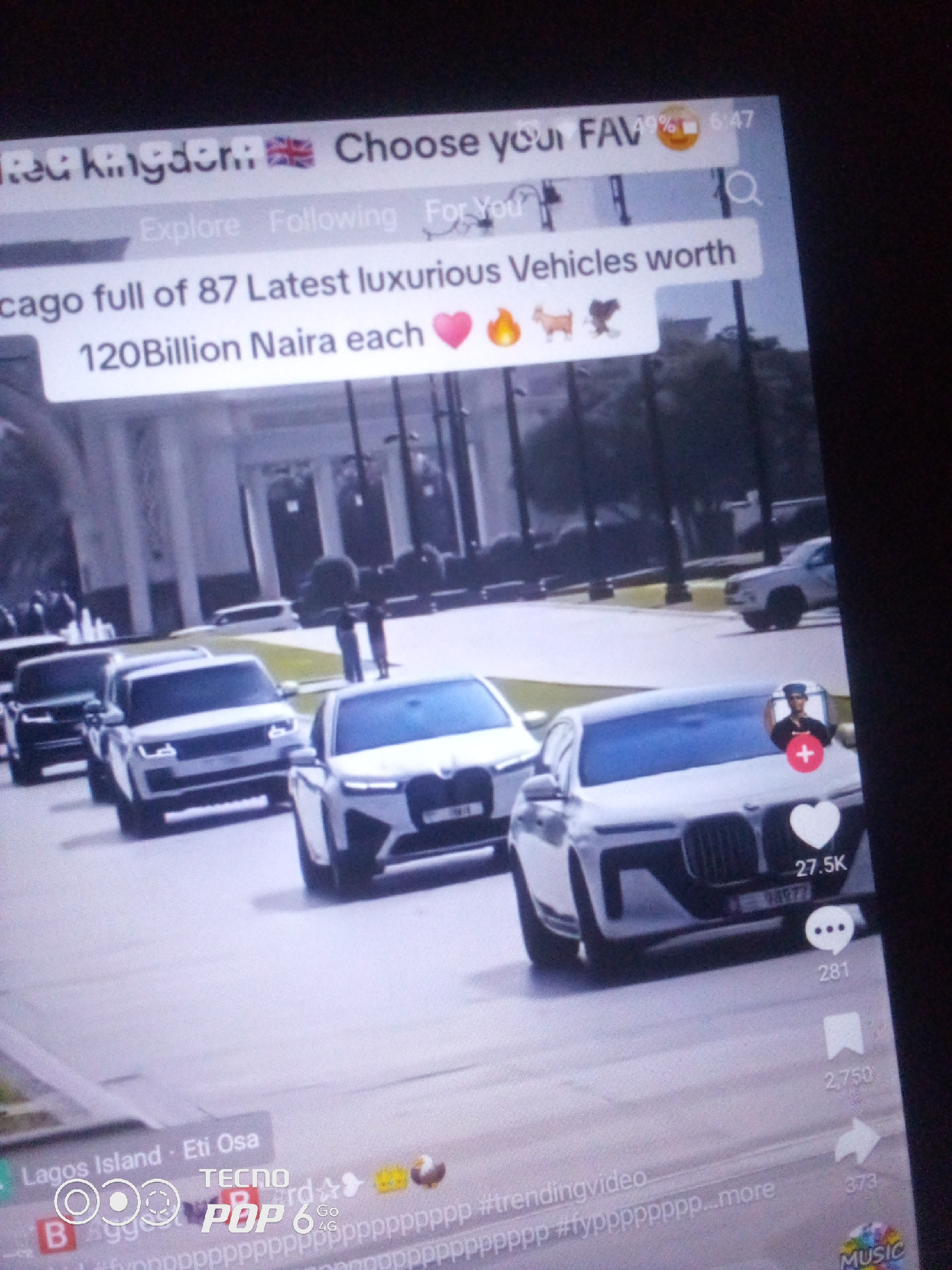 The image displays a TikTok-like video interface overlaid on a live-action scene. The video content appears to be a compilation of luxury vehicles, possibly in a procession or parade. Text overlays on the video state "chicago full of 87 Latest luxurious Vehicles worth 120Billion Naira each" accompanied by emojis of a heart, fire, and animals. A search bar is visible at the top with "Choose your FAV" and a UK flag emoji. The bottom of the screen shows video metadata including "Lagos Island - Eti Osa" and "TECNO POP 6 Go" with hashtags and interaction counts for likes, comments, and shares. The background of the video depicts a street with modern buildings and trees, suggesting an urban setting. The screen itself appears to be a mobile phone, held at an angle, with the surrounding environment being dark.