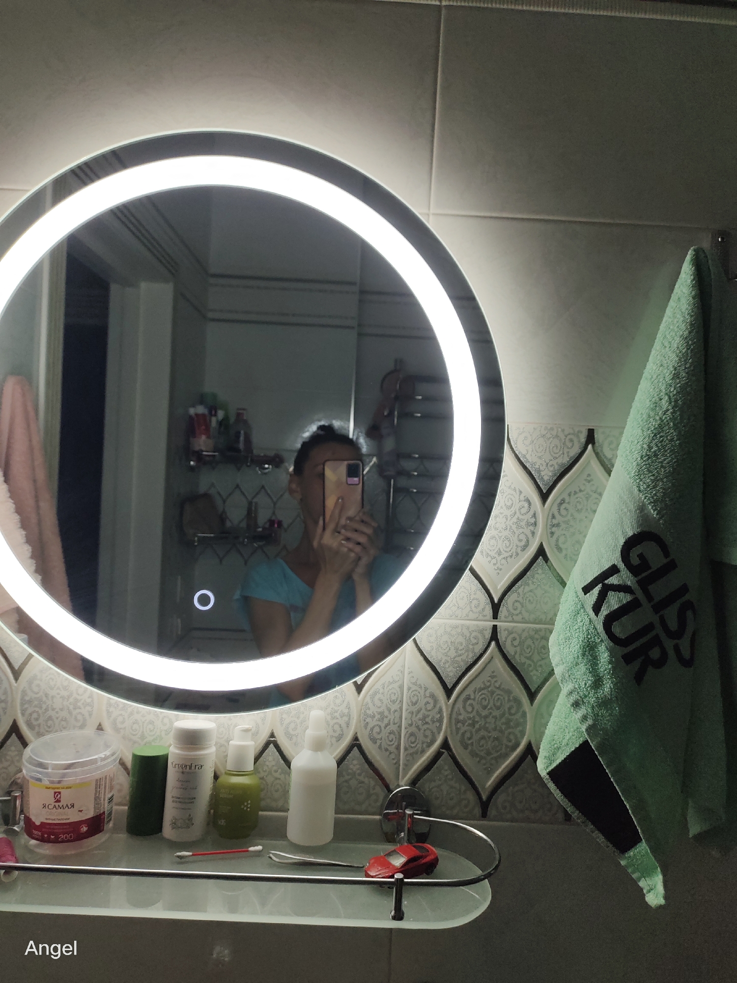 The image depicts a bathroom scene, likely taken in Tula, Russia, given the text "Angel" in Russian at the bottom of the frame. A circular mirror with an integrated LED light is the central focus. A woman is visible in the reflection, holding a smartphone and taking a selfie. On a shelf below the mirror, several toiletries are arranged, including a container labeled "Я САМАЯ ORIGINAL," a green bottle, a bottle labeled "GreenFra," a green pump bottle, and a white bottle. A small red toy car is placed on a rack to the right of the shelf. A light green towel with the text "GLISS KUR" is hanging on the right side of the frame. The background features tiled walls.