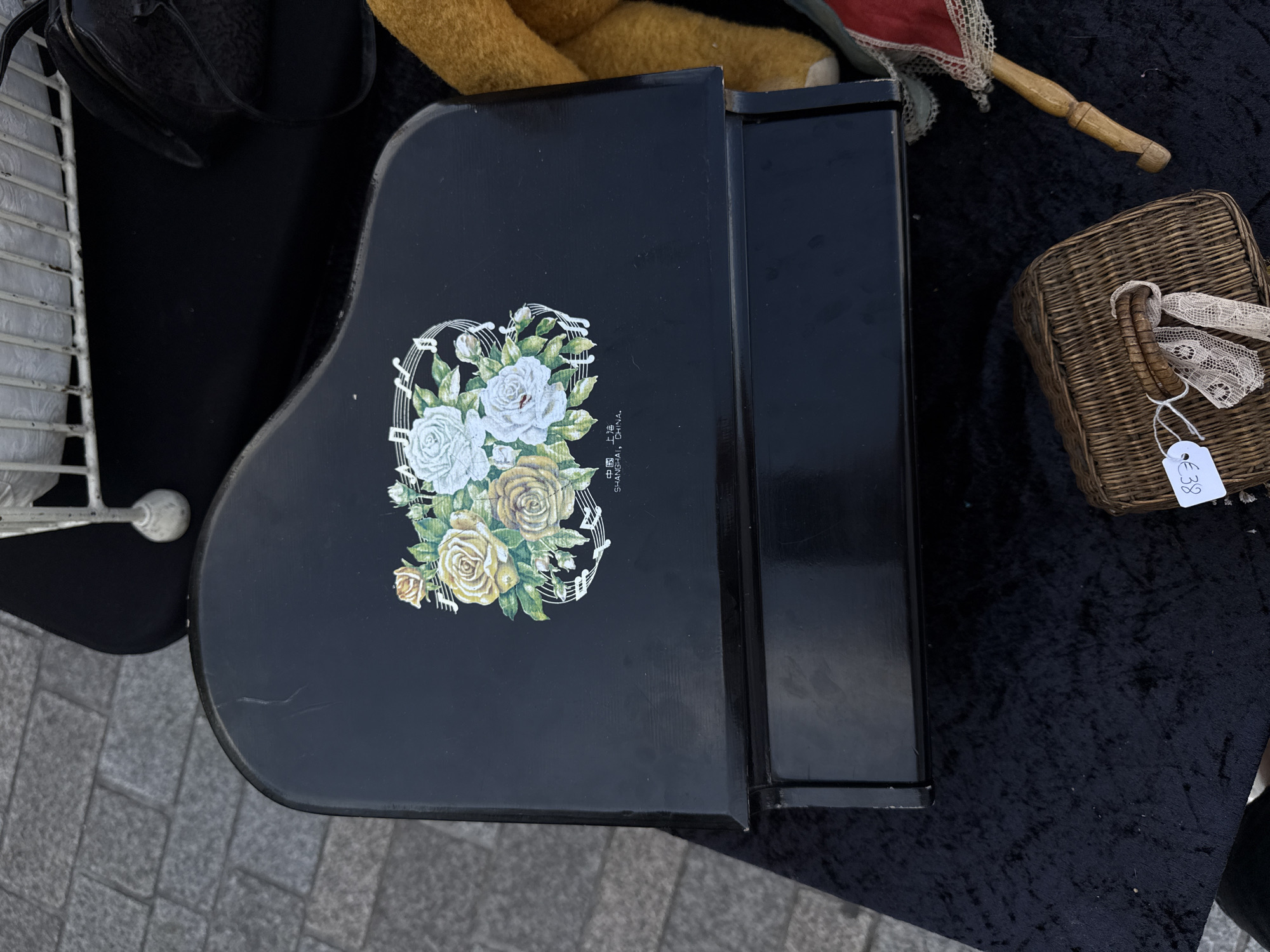 A high-angle, eye-level shot shows a black, toy grand piano with a floral decal on its lid, resting on a black velvet cloth. The piano is adorned with a bouquet of yellow and white roses, interspersed with musical notes. Beneath the roses, Chinese characters and the text "SHANGHAI, CHINA" are visible. To the right of the piano, a portion of a yellow teddy bear and a red and white fan are visible. In the lower foreground, a small wicker basket with a lace ribbon tied around its handle is placed on the velvet cloth. A white price tag with the inscription "€38" is attached to the basket. The scene appears to be outdoors, with a glimpse of a grey cobblestone ground in the upper left corner. The overall setting suggests an outdoor market or flea market stall, possibly in the city of Bruges, Belgium, given the historical cobblestone street visible.